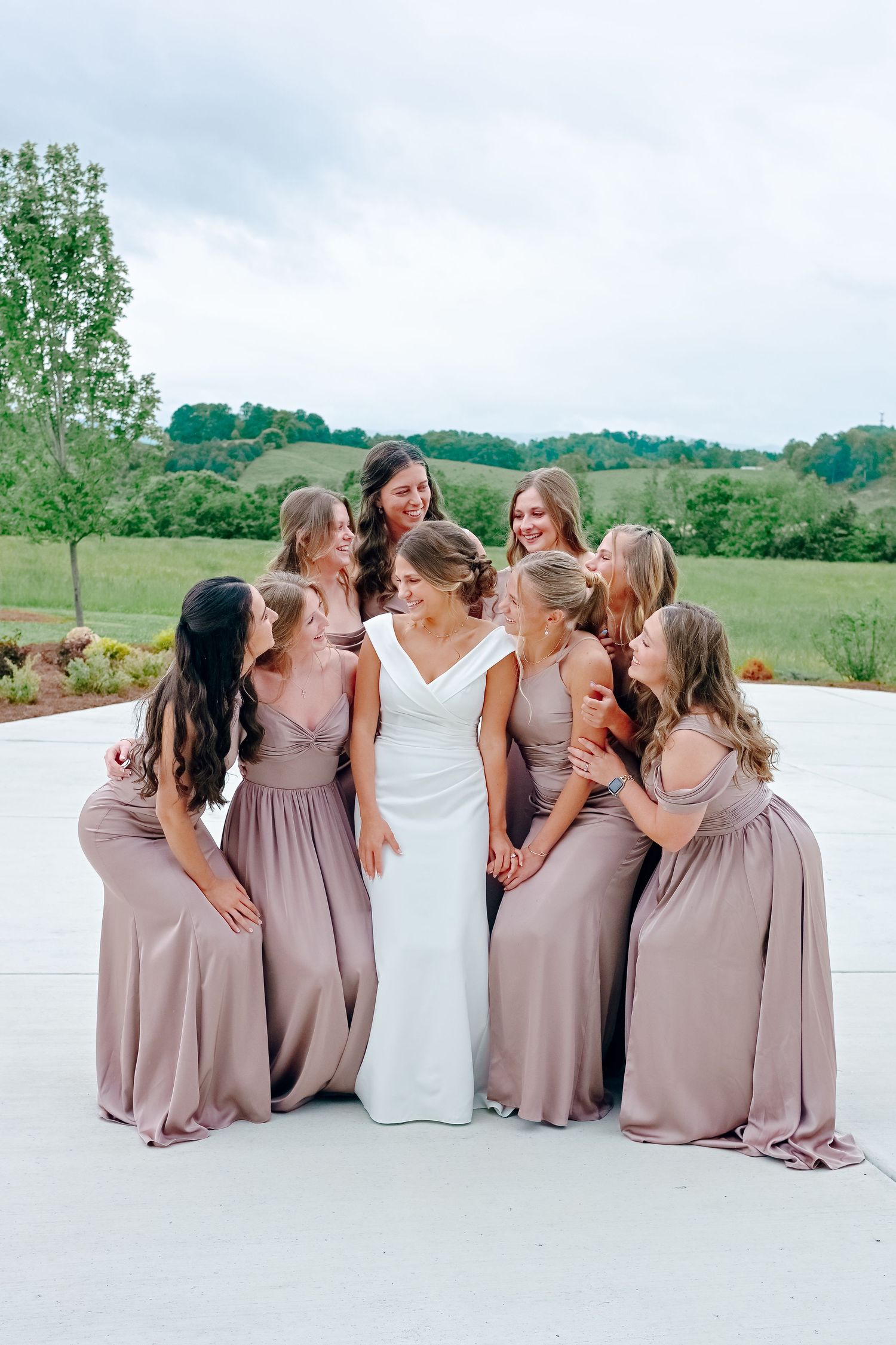 Wedding party in blush dresses sits together on white surface for casual group photo.