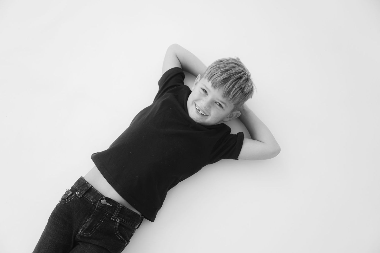 Black and white photo of a smiling boy lying on his back with hands behind his head, wearing a black t-shirt and jeans.