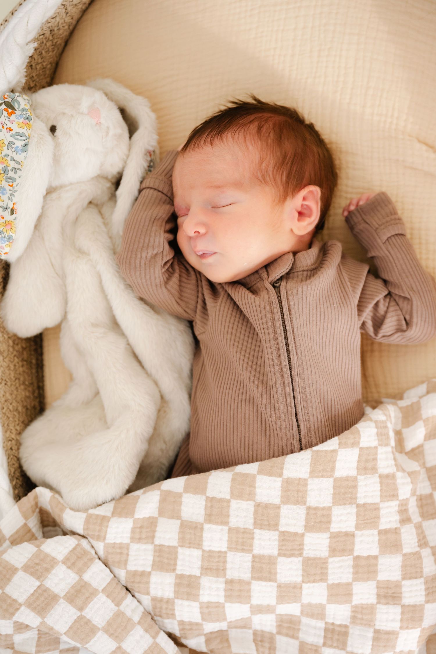 Newborn baby sleeping peacefully in a bassinet wearing a brown zip-up outfit, with a white bunny stuffed animal nearby.