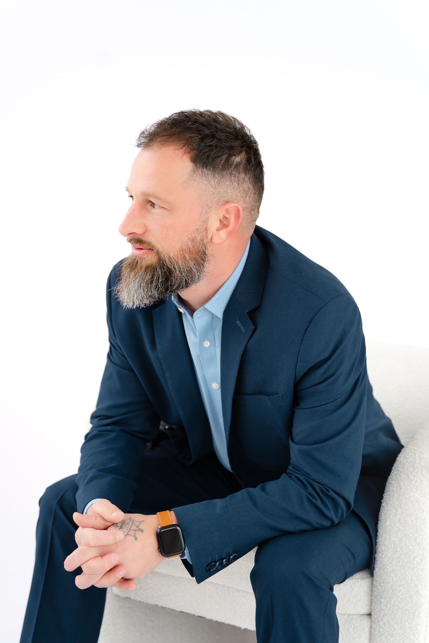 Professional in navy suit and light blue shirt sits thoughtfully against white background in multi-angle portrait series.