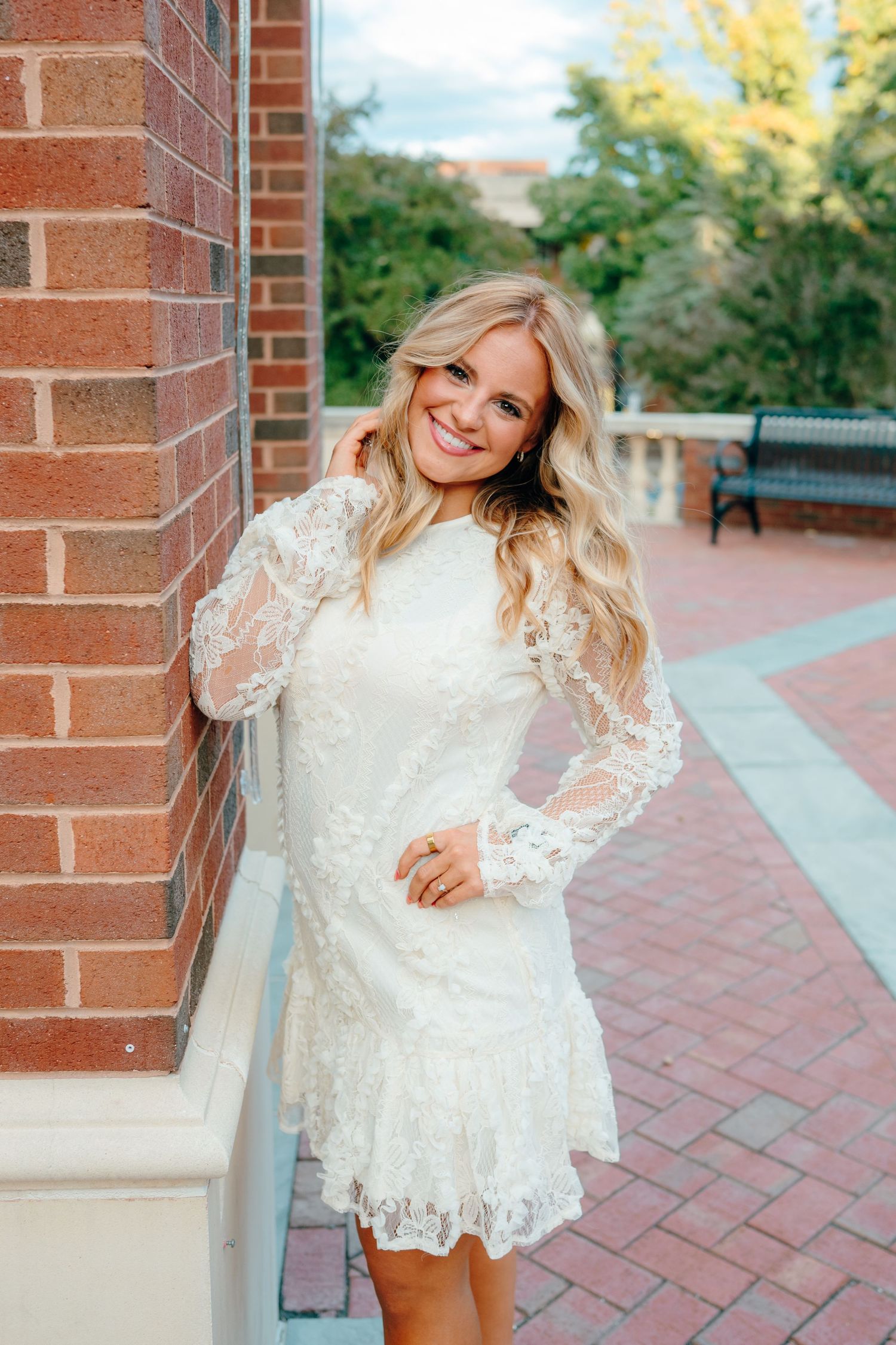 Graduate in white lace dress poses against brick wall on campus.