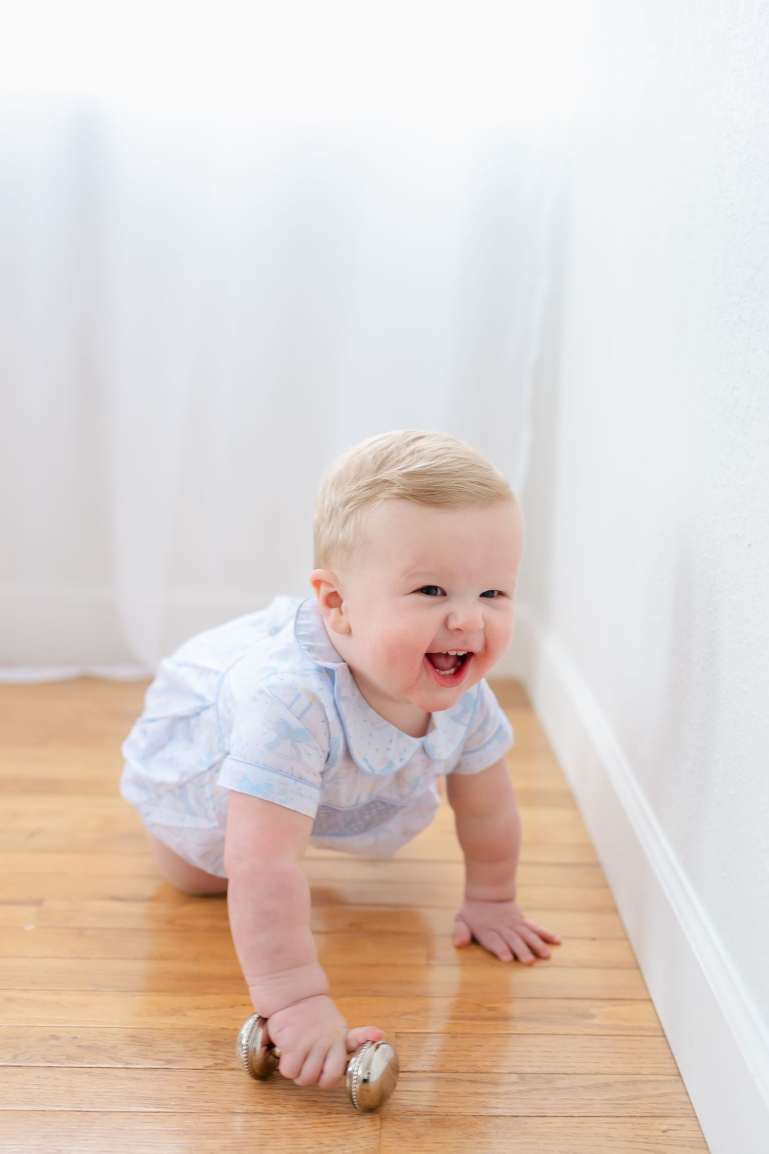 A baby in a white outfit crawls happily across a hardwood floor near a white baseboard.