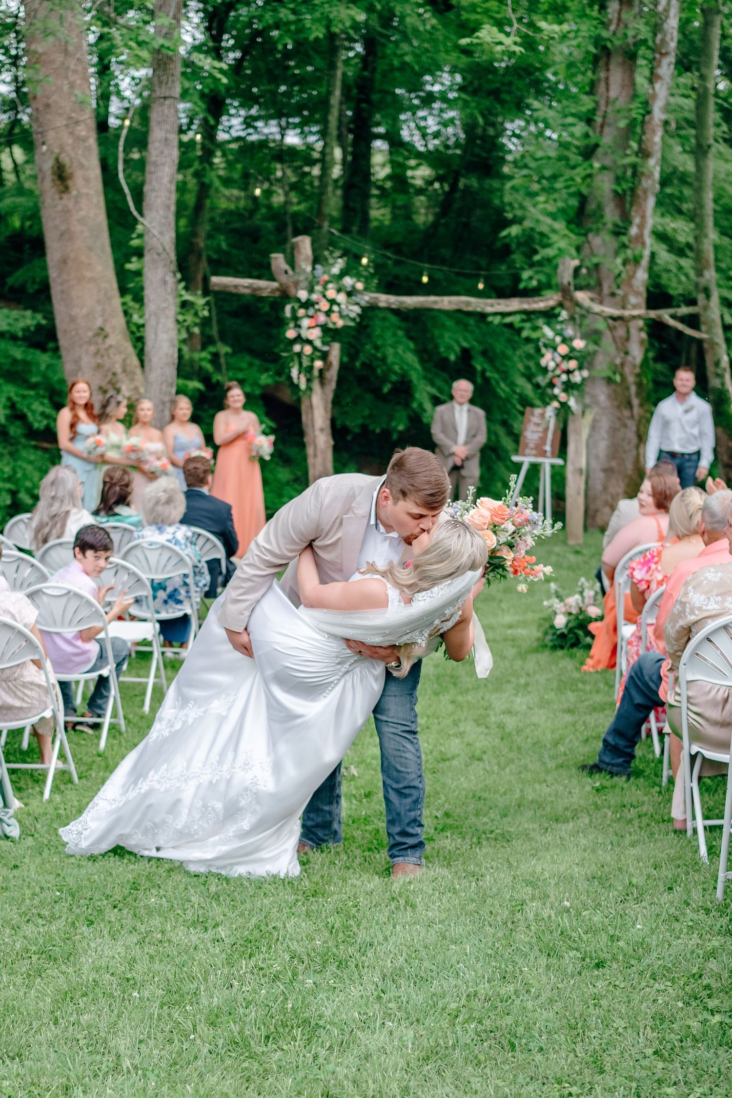 Outdoor woodland wedding ceremony with floral arch and white chairs on green lawn.