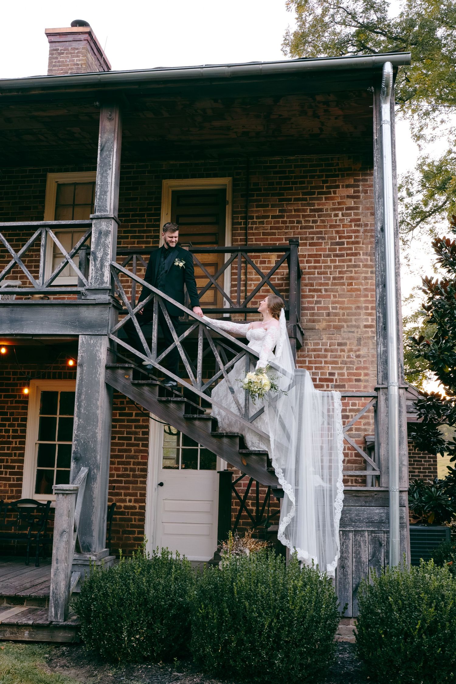 Bride and groom hold hands on rustic brick building staircase, bride in flowing white gown with veil and bouquet.