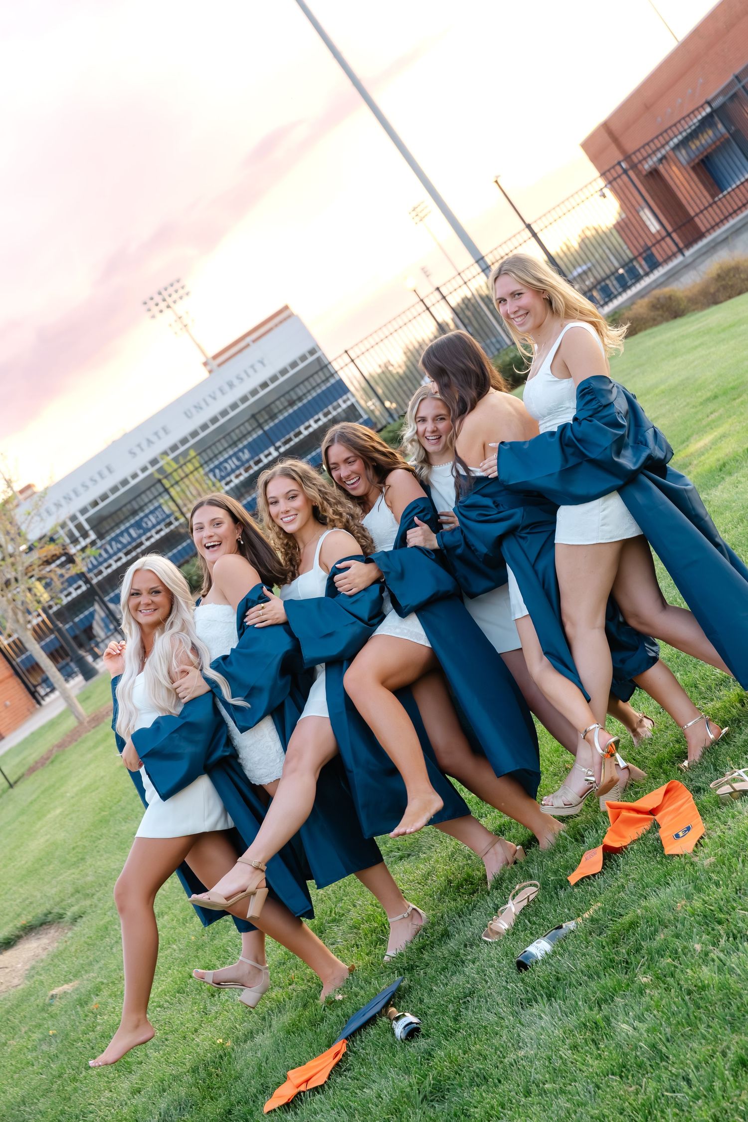 A group of young women in white dresses celebrate graduation outdoors, holding teal caps and gowns on a green field.