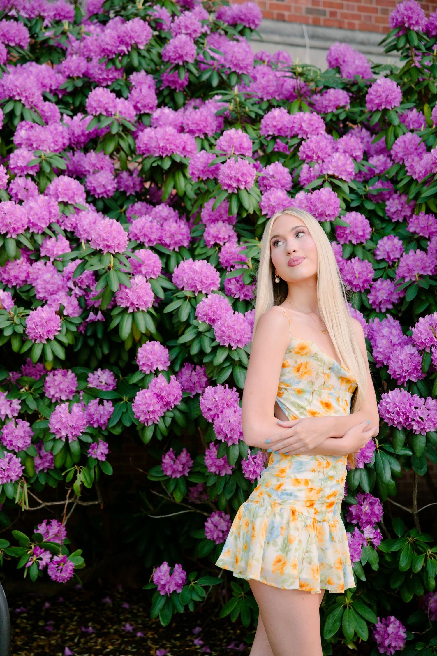 Young blonde woman in floral yellow dress poses before vibrant purple rhododendron blooms in lush garden setting.