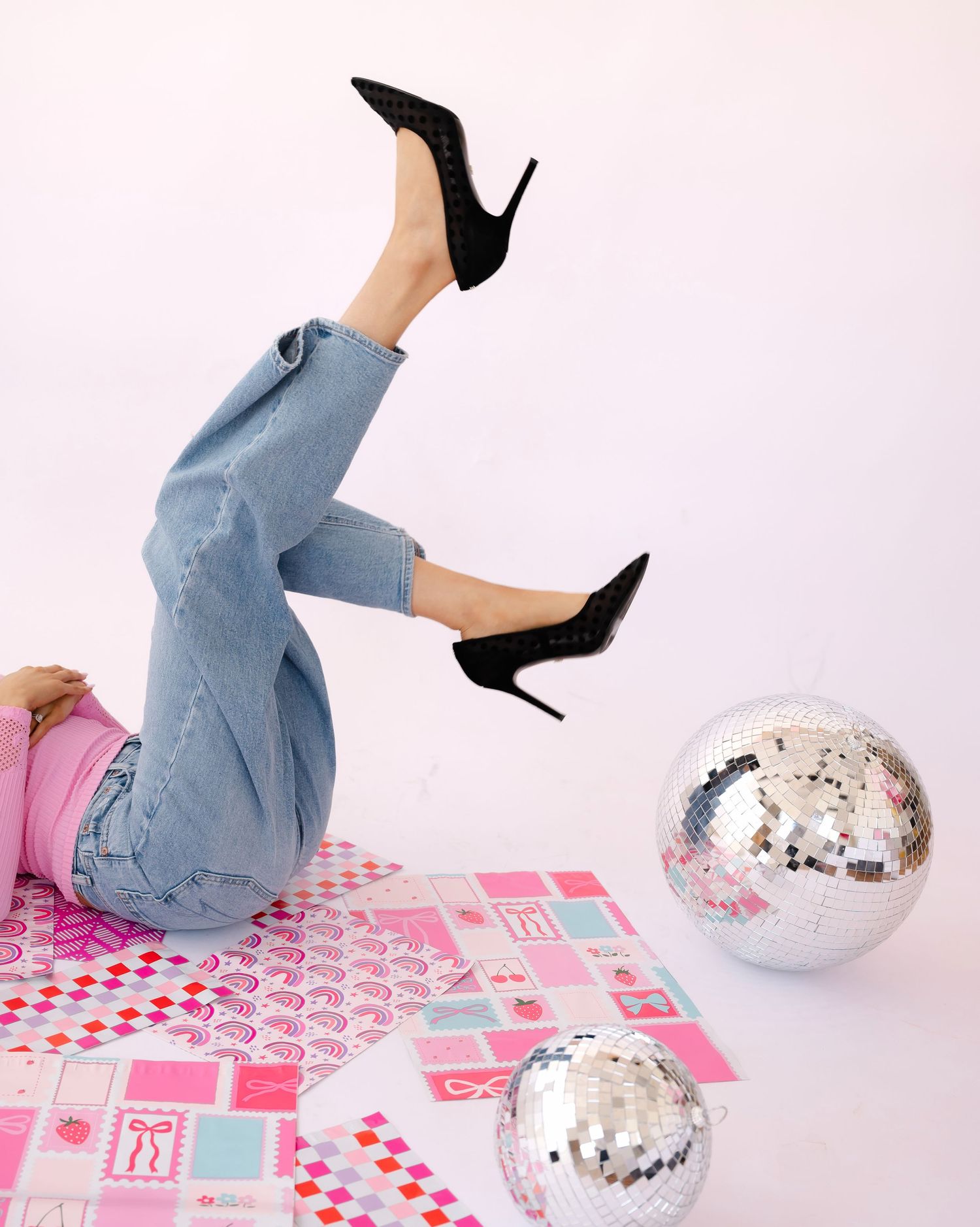 Playful leg poses with black stilettos and blue jeans against pink geometric floor with disco balls.