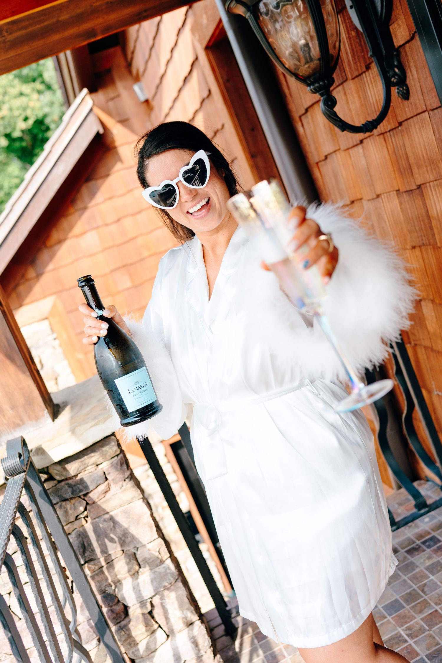 Bride in white dress and sunglasses poses playfully in front of rustic wooden backdrop.