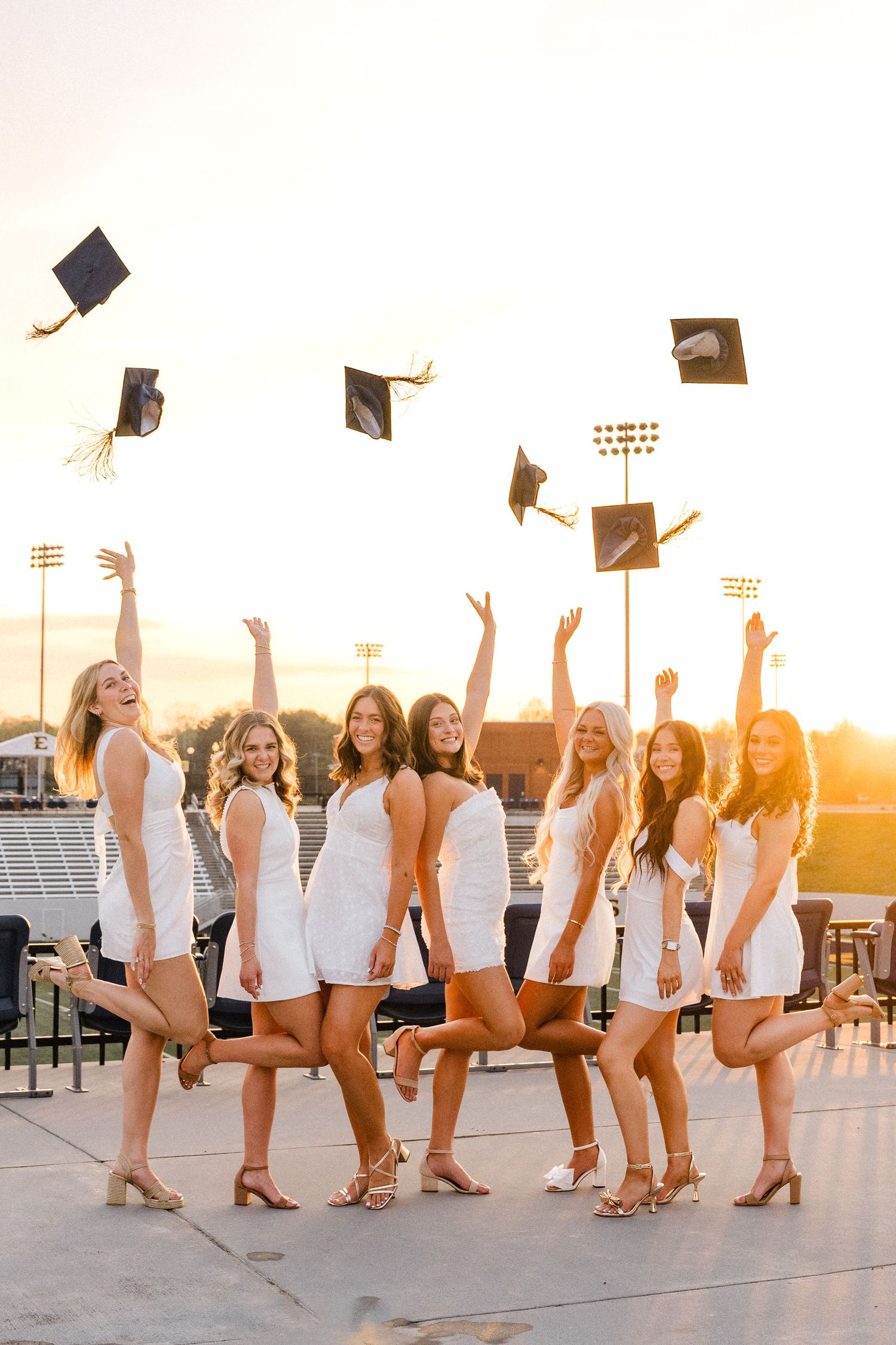Seven women in white dresses toss graduation caps in the air at sunset on a stadium rooftop.