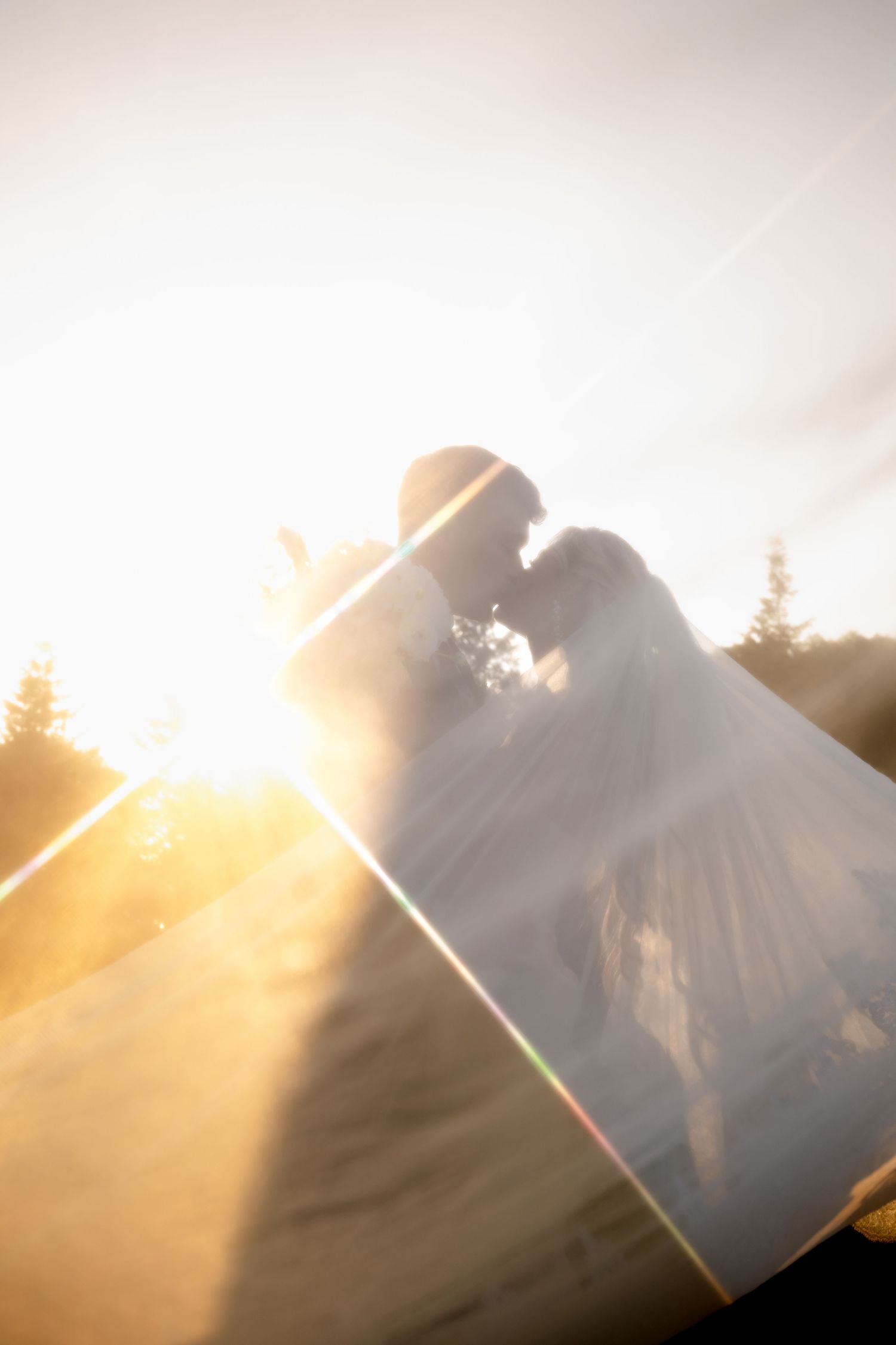 Bride and groom silhouetted against golden sunset light, sharing a romantic kiss with lens flare and veil flowing.