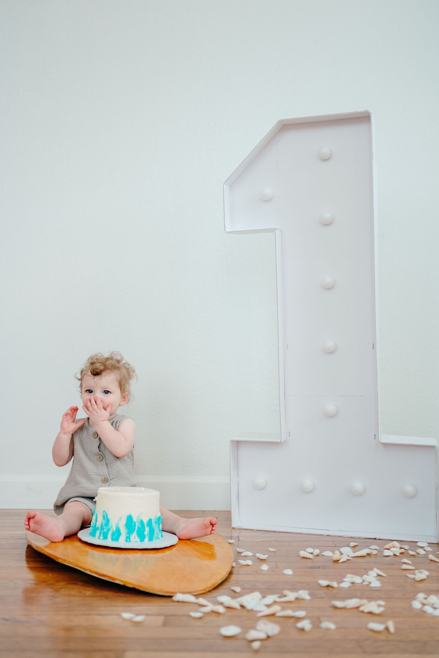A baby sits on a round cushion with a blue and white cake, surrounded by scattered pieces of paper on a wooden floor.