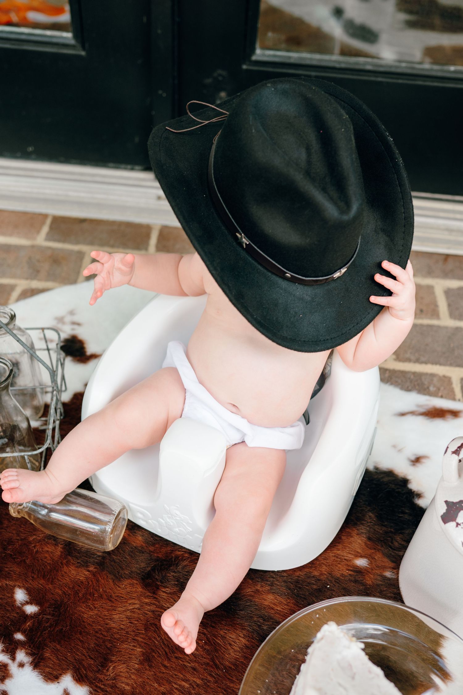 A baby in a black cowboy hat sits on a brown cowhide rug in a series of candid photos.