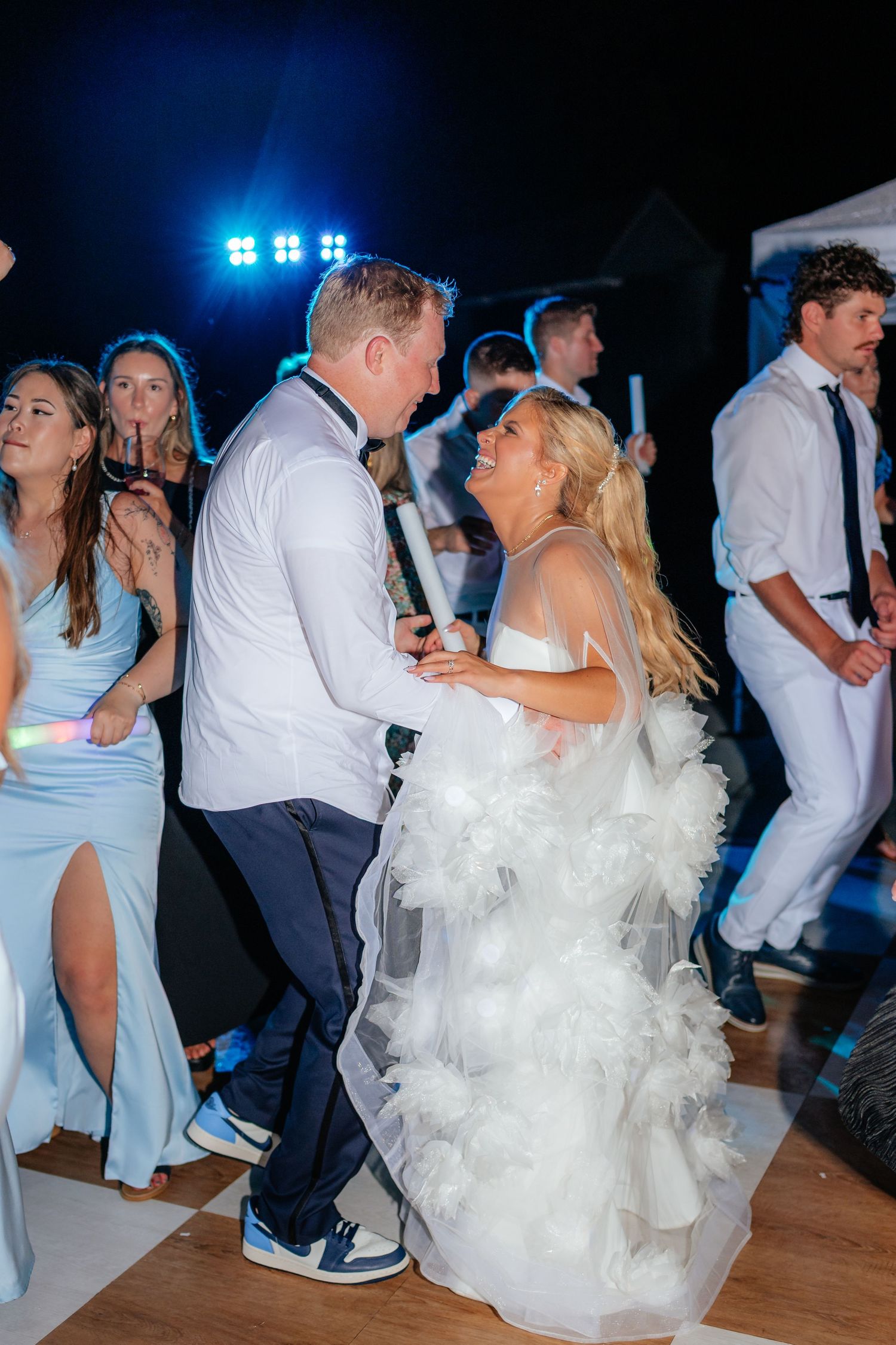 Wedding guests dancing on illuminated dance floor with bride in white gown.