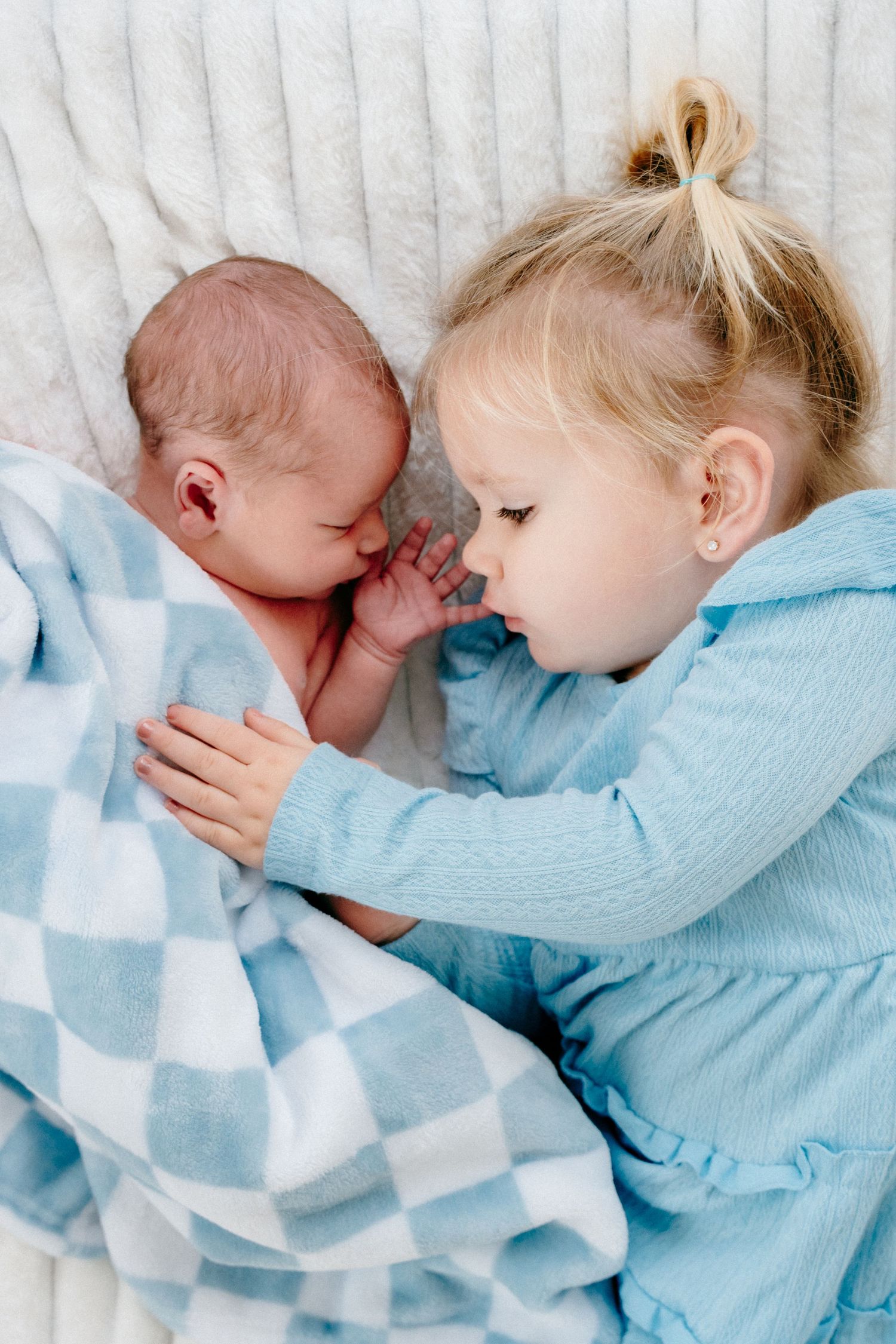 Siblings in blue outfits share tender moment while lying on white blanket.