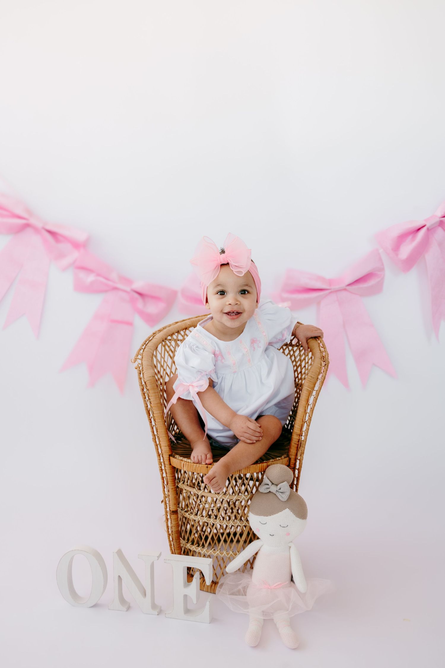 Baby sitting in wicker chair with pink bow decorations and white letters.