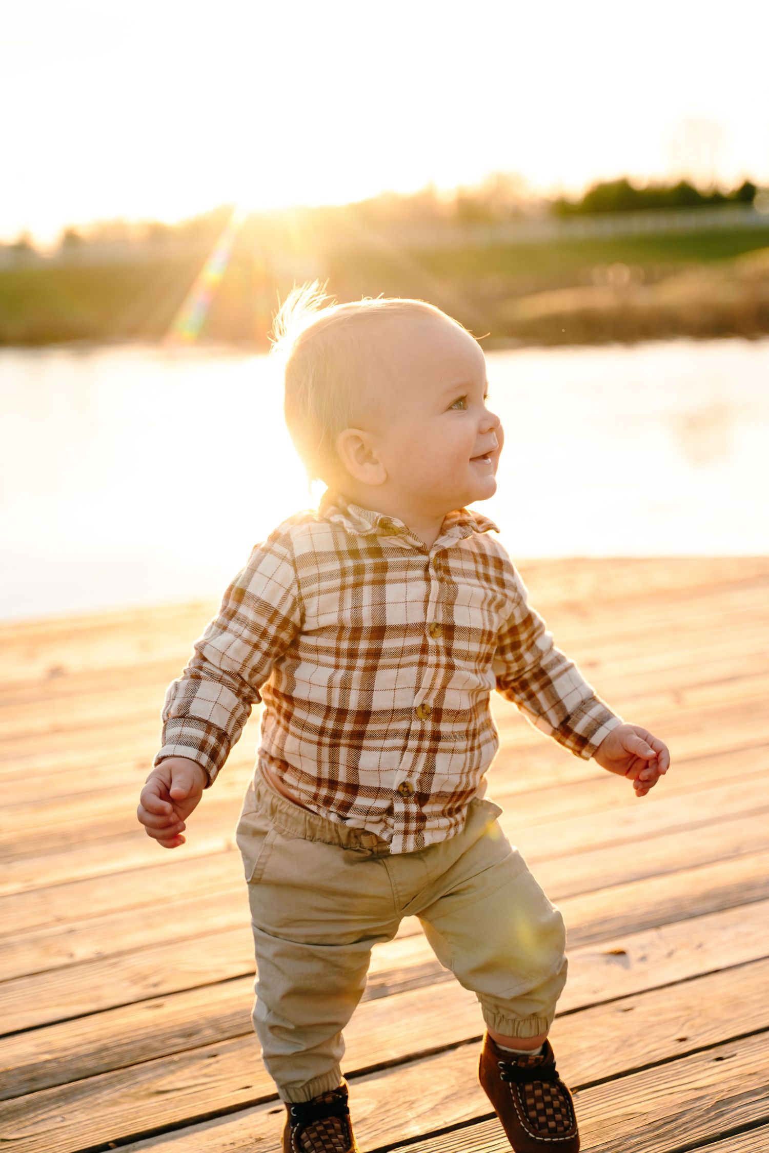 Baby in plaid shirt walks on wooden deck during golden sunset.