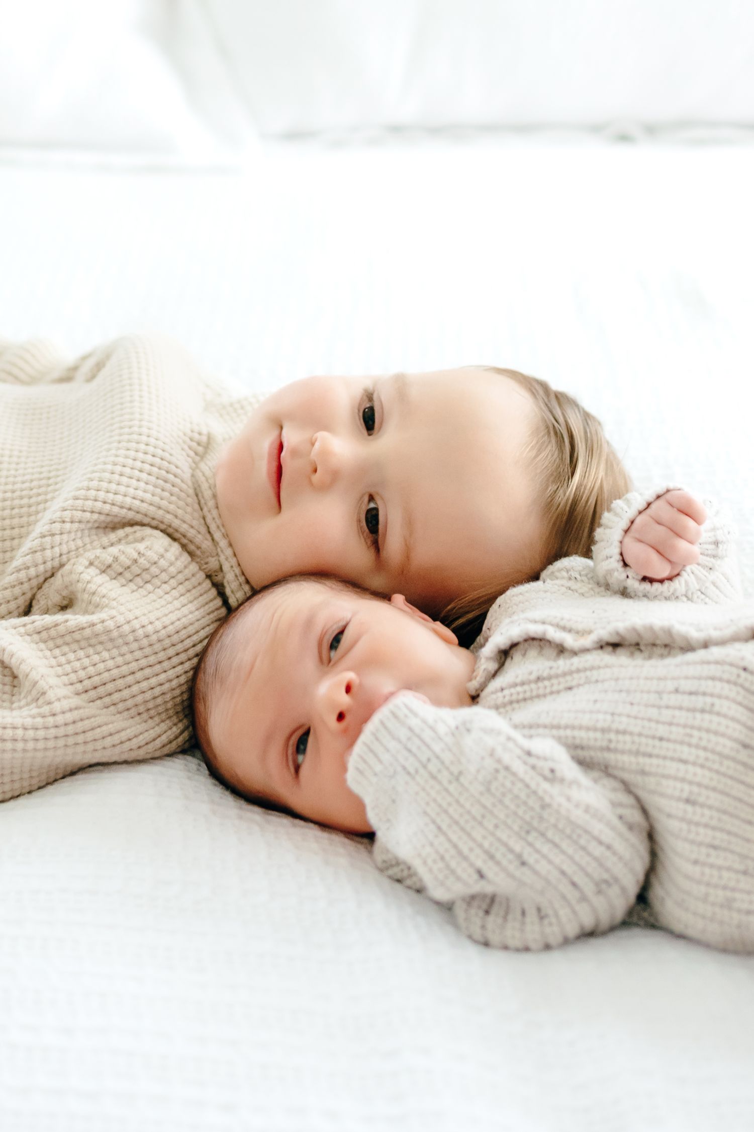 Two siblings in matching knit sweaters cuddle on white bedding.