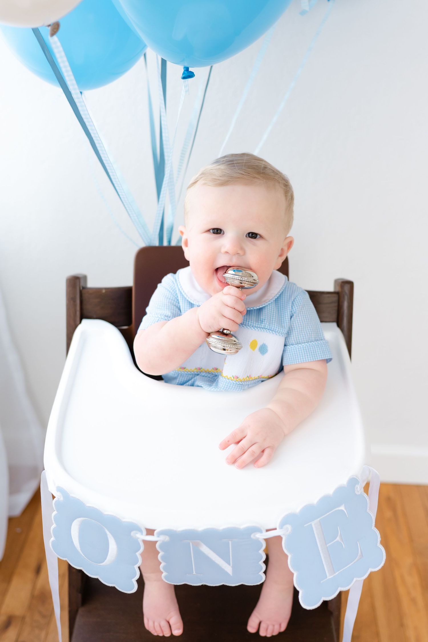 A baby in a high chair with light blue balloons and a 'ONE' banner celebrating their first birthday milestone.