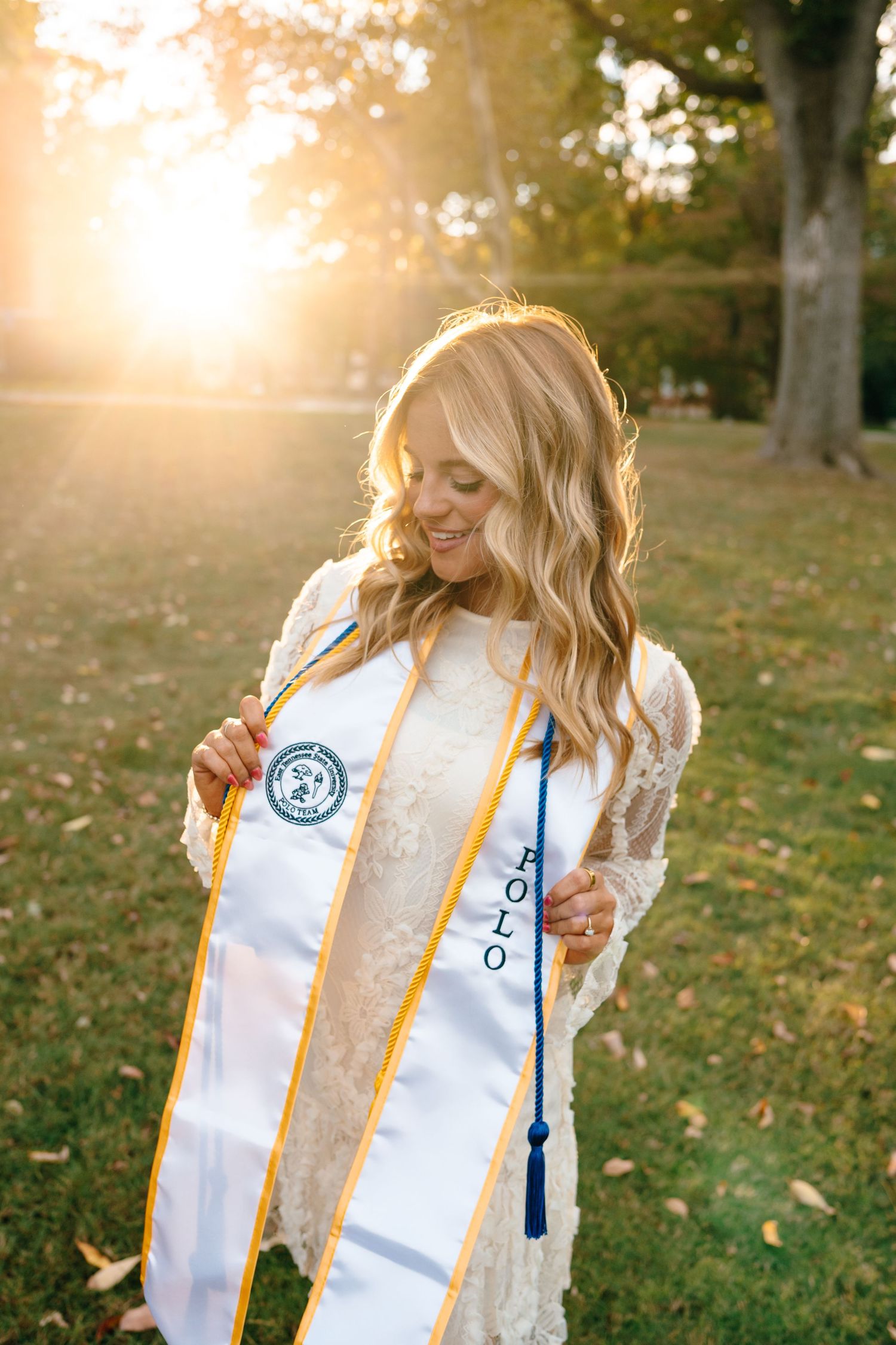 Graduate in white dress displays honor stole during sunset photoshoot.