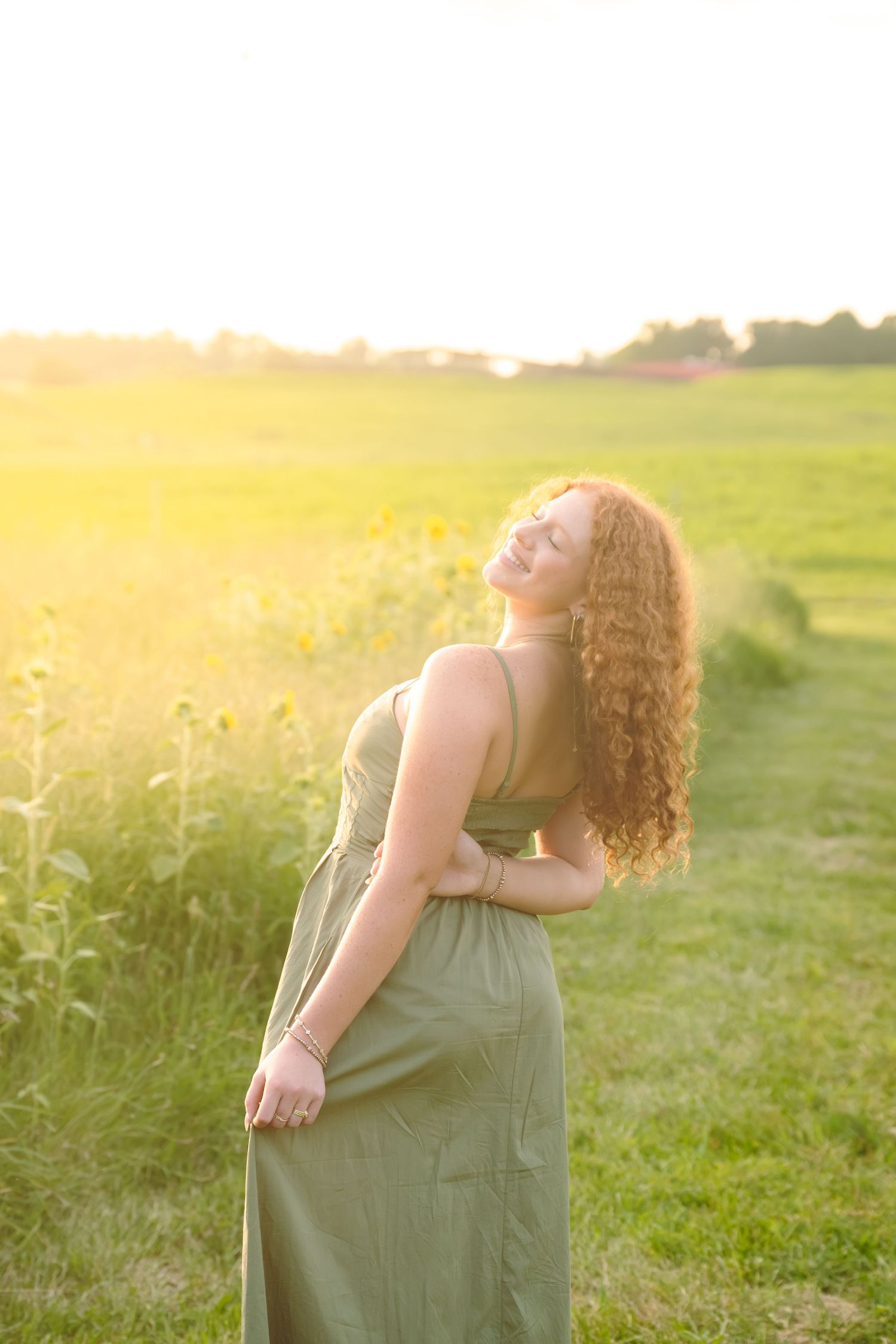 Young woman with curly hair in olive green maxi dress posing in sunflower field at golden hour sunset.
