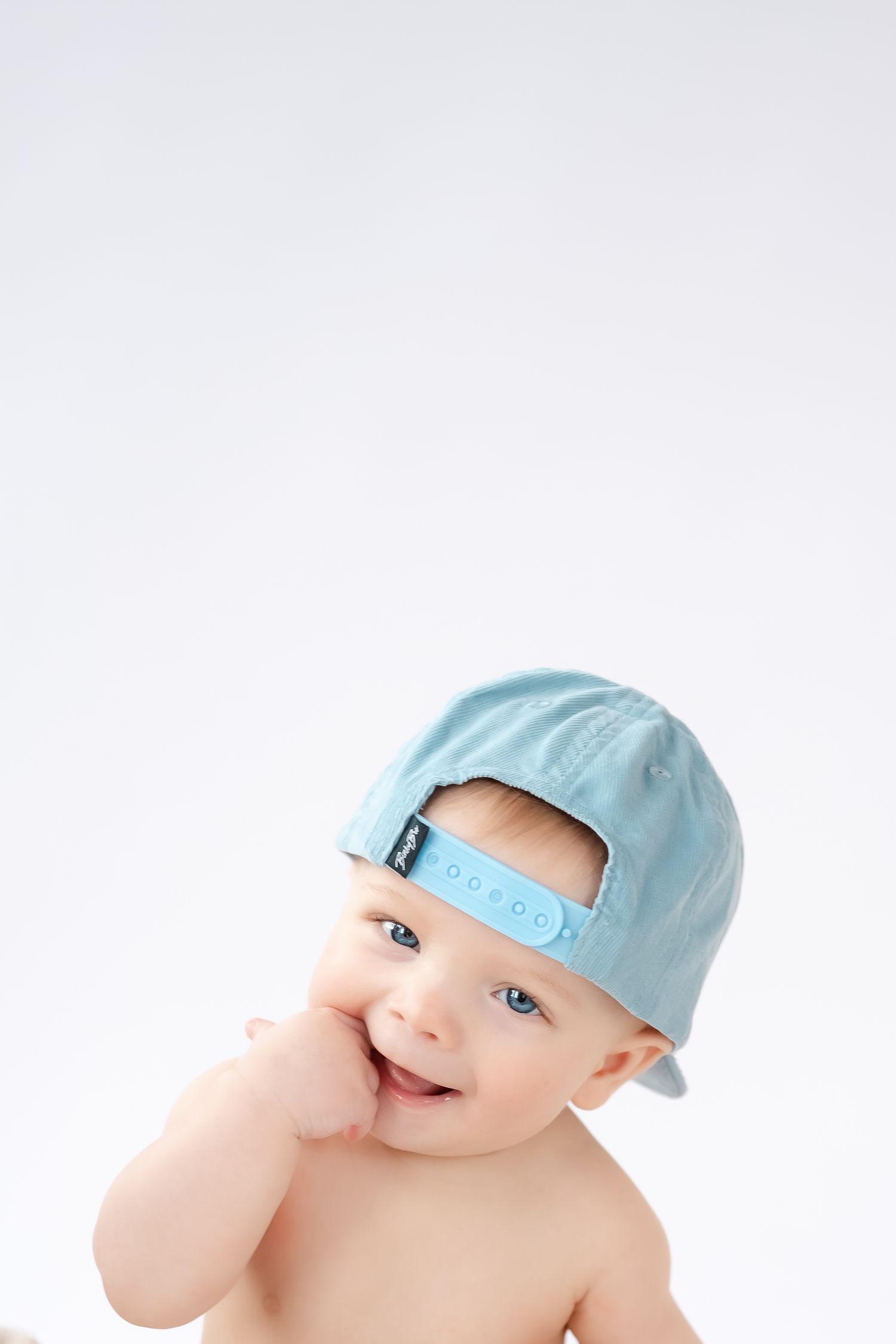 Baby wearing light blue baseball cap smiles while playing peek-a-boo against white background.