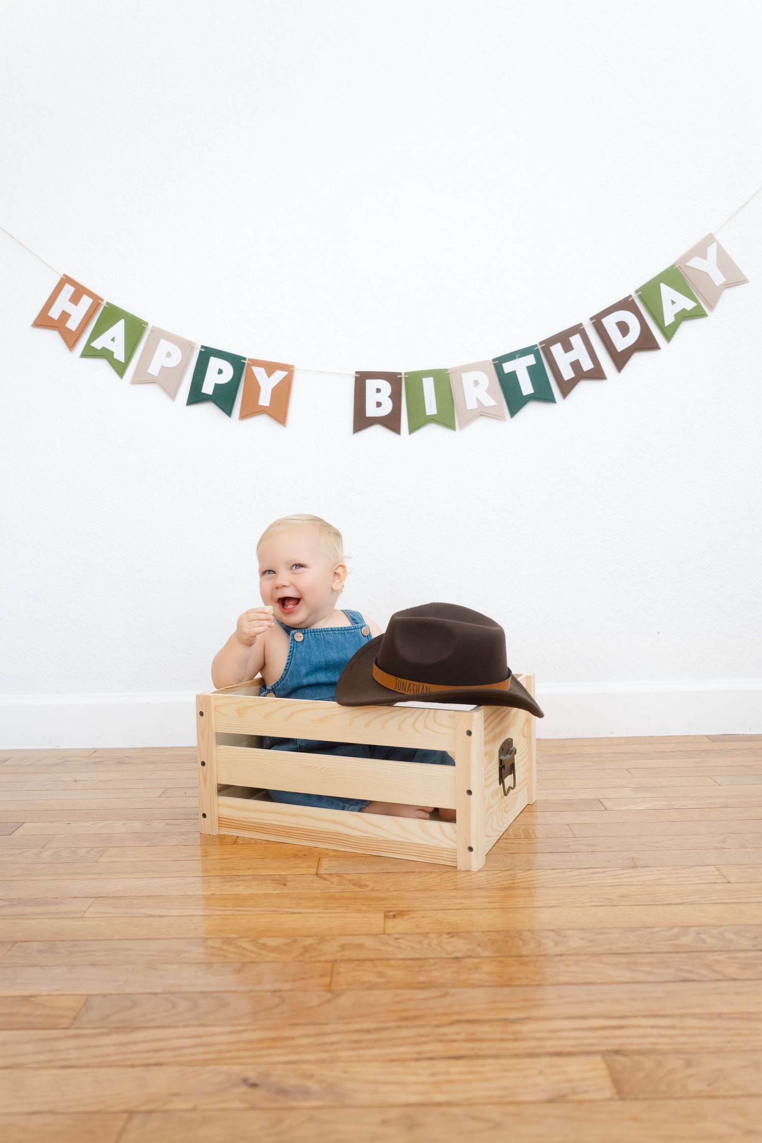 Happy baby in wooden crate with cowboy hat under colorful Happy Birthday banner on white wall.