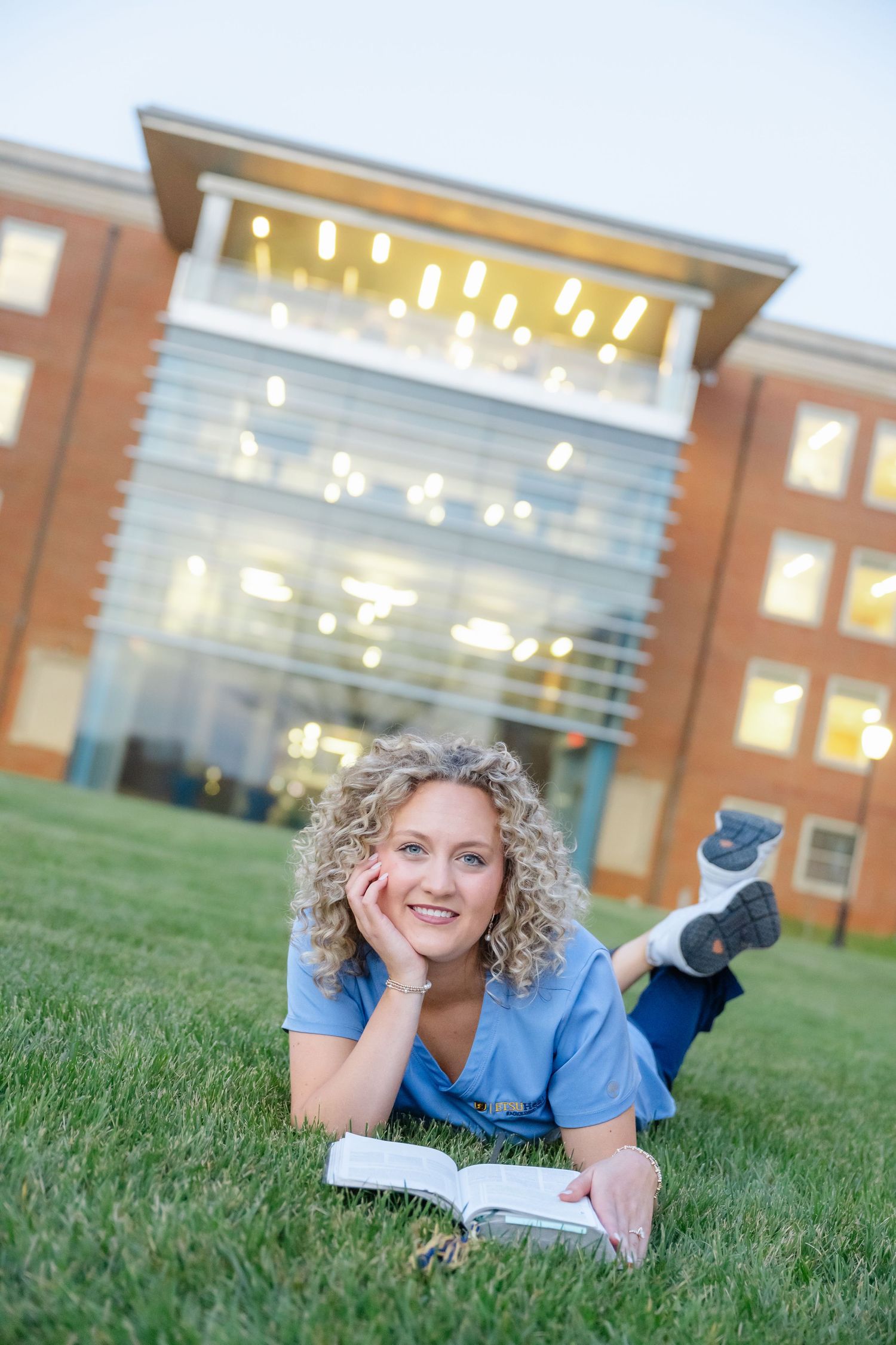 A smiling nursing student in blue scrubs lies on green grass reading a textbook outside a modern brick university building.