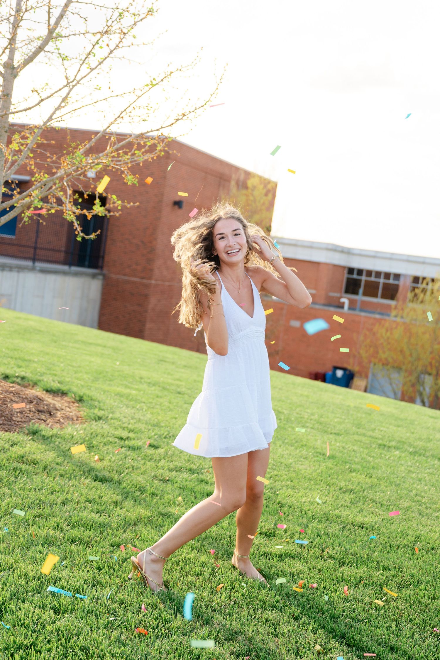 Young woman in white dress smiling and dancing outdoors with colorful confetti falling on green grass.