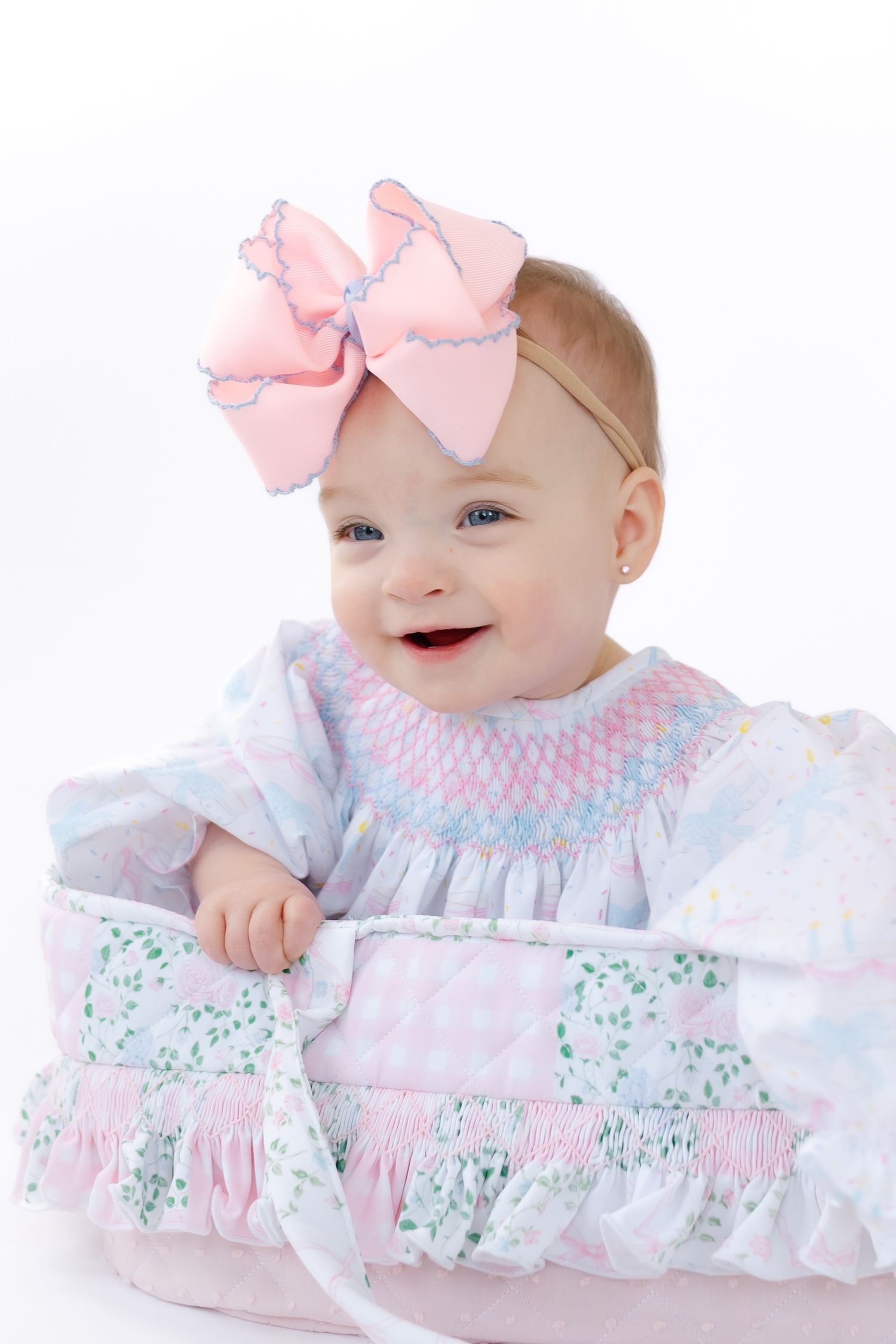 Baby wearing white and pink smocked dress with floral print and pink bow headband lying on white background.
