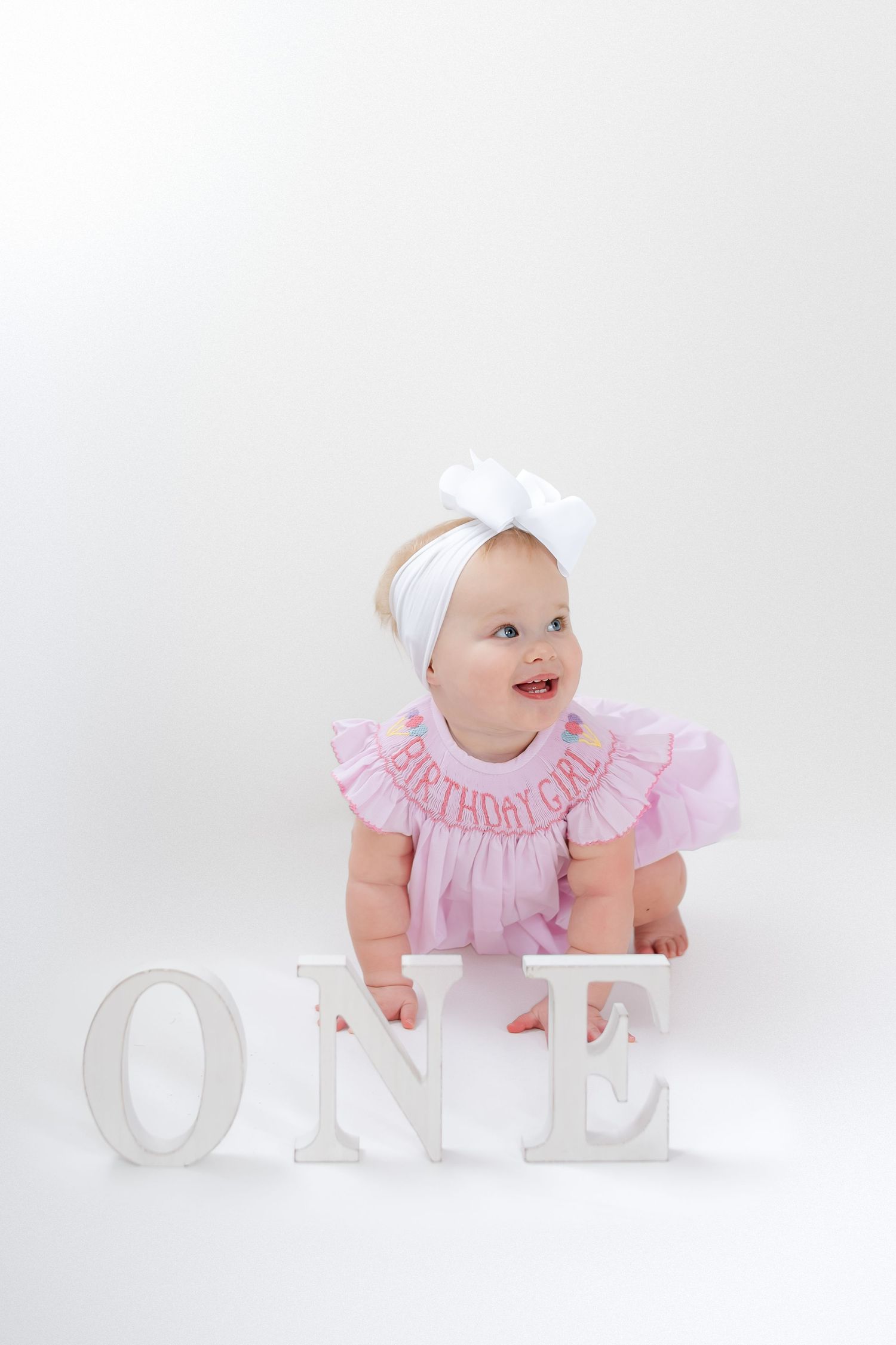 Baby in pink ruffled dress crawls beside white decorative letters spelling 'ONE' against light background.