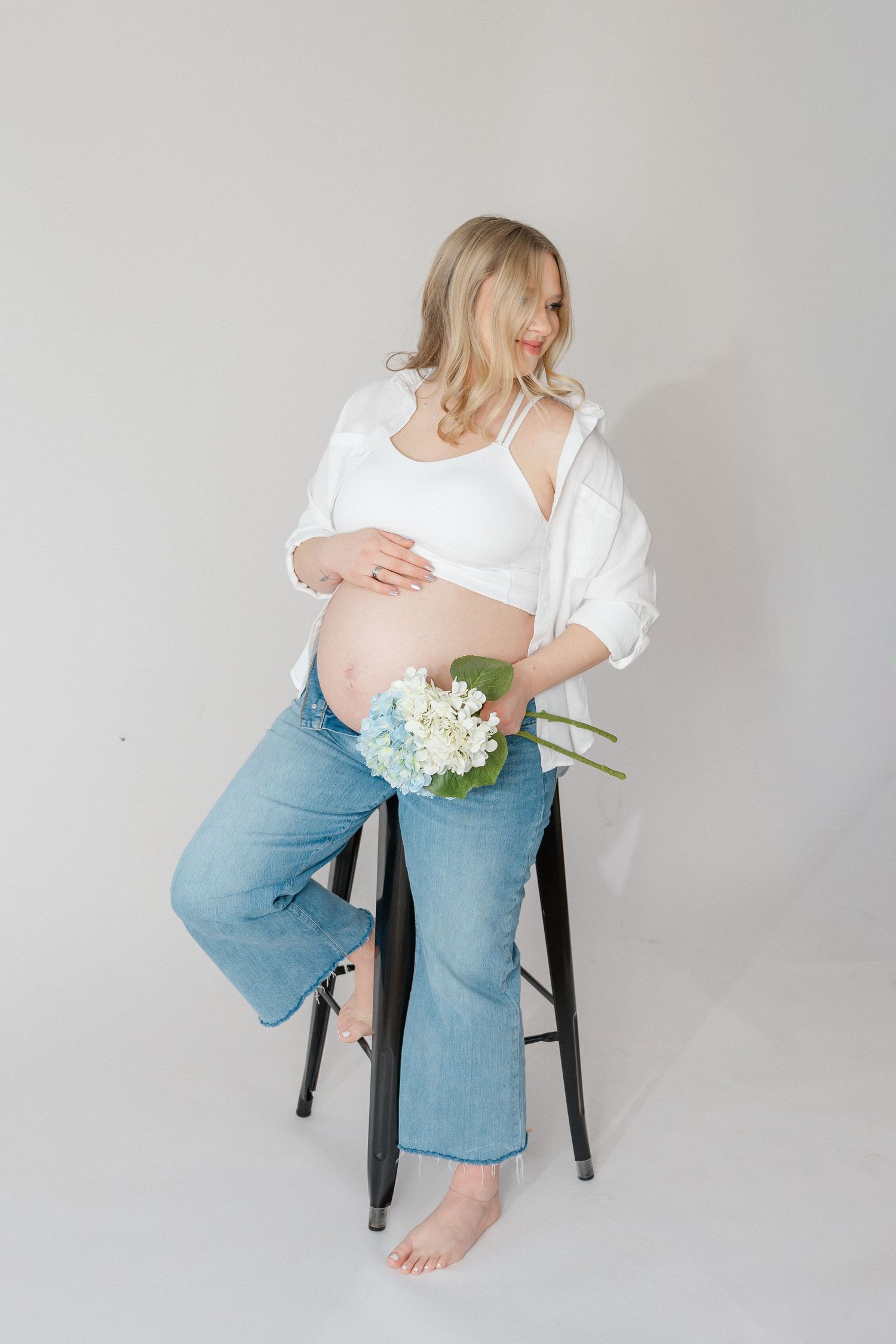 Pregnant woman in white top and jeans sits on stool holding white flowers in minimalist maternity photo.