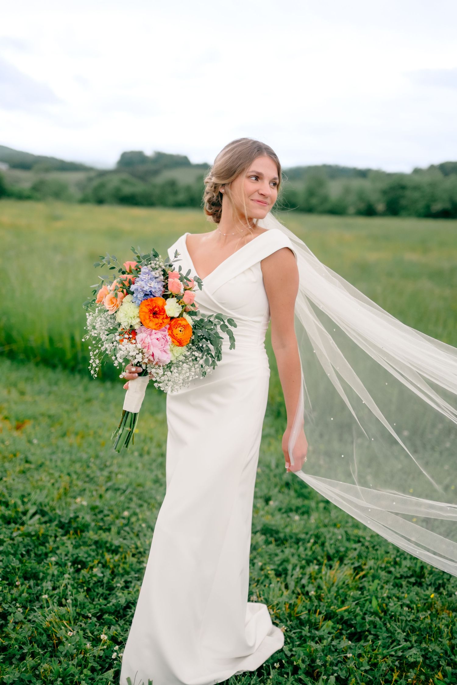 Bride in elegant white dress holds colorful bouquet with flowing veil in meadow.