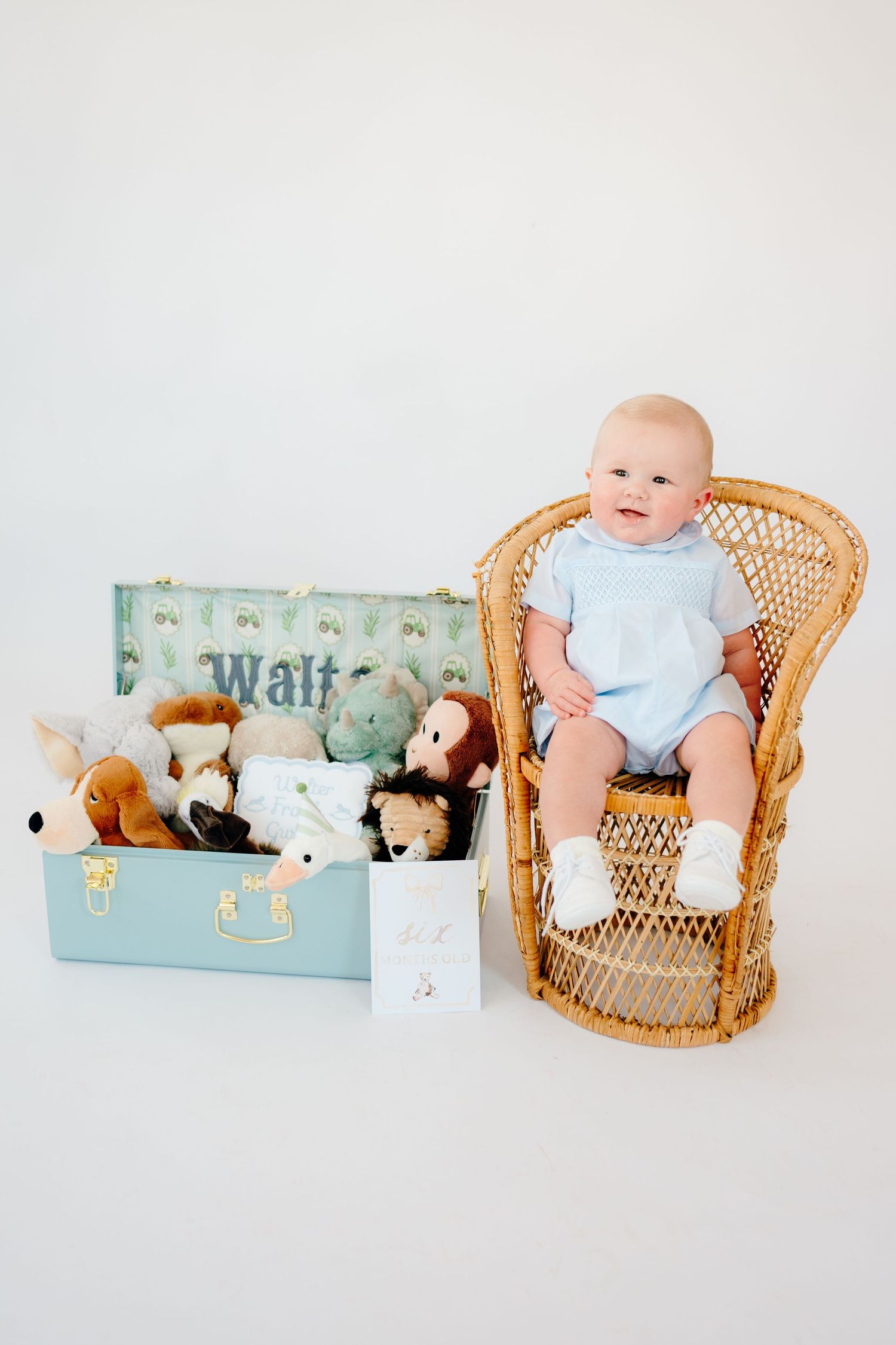 Baby in blue outfit sits in wicker chair next to vintage suitcase filled with stuffed animals.