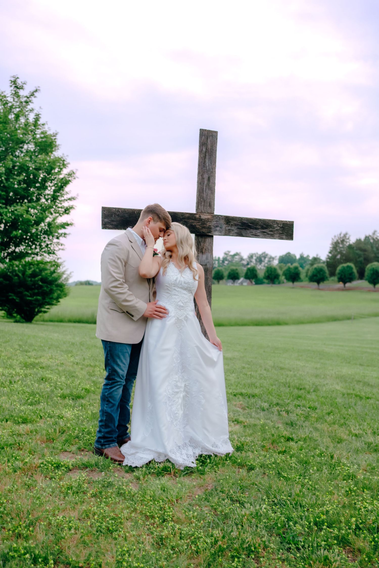 Wedding couple shares intimate moment near rustic wooden cross in countryside setting.