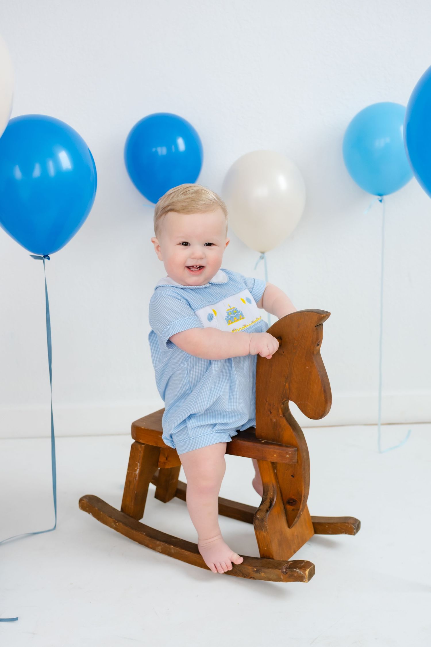 Baby in blue romper plays with wooden rocking horse toy against white backdrop with blue balloons.