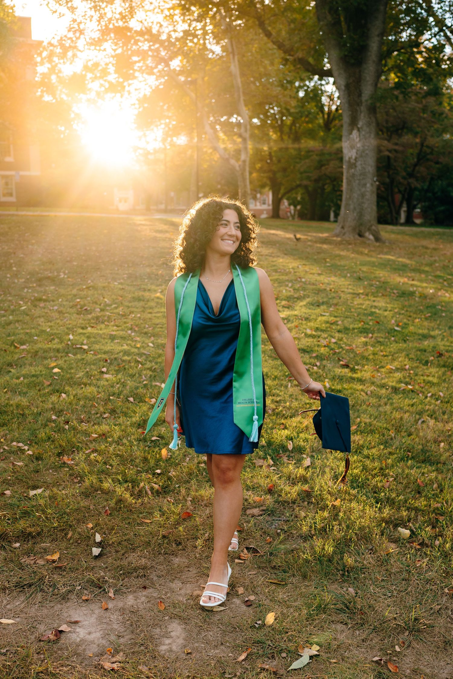 Graduate in navy dress and green stole walks through sunny park during golden hour.