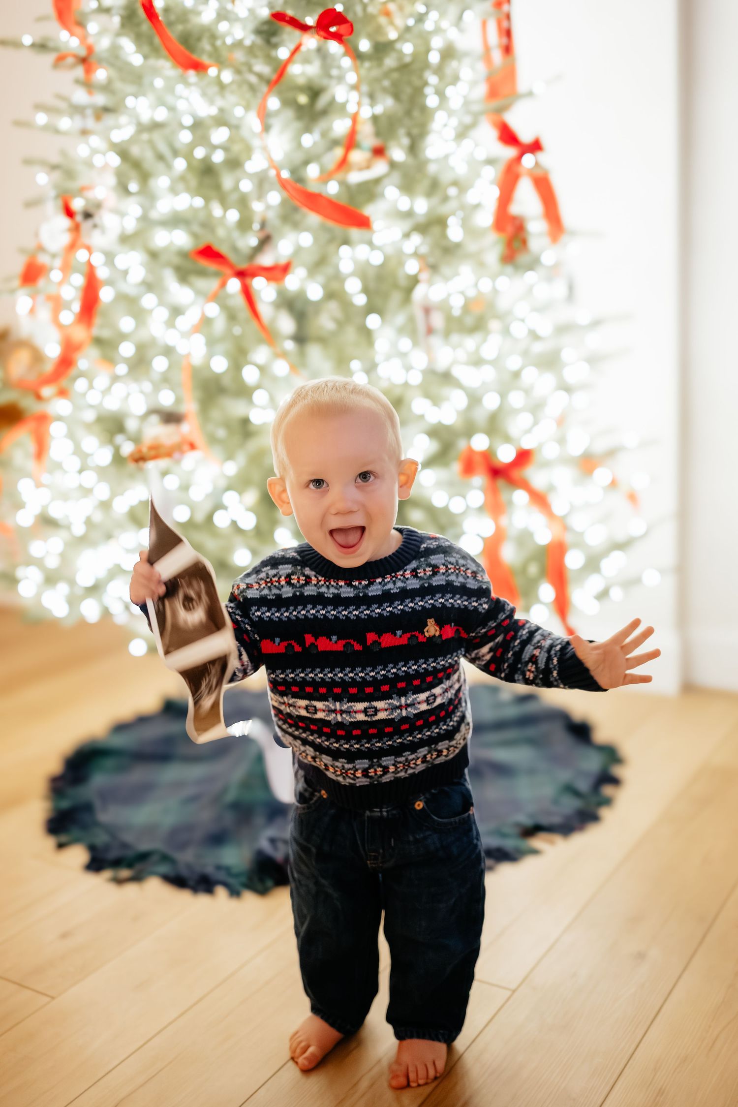Baby in festive sweater crawling by Christmas tree with red ribbons and decorations.