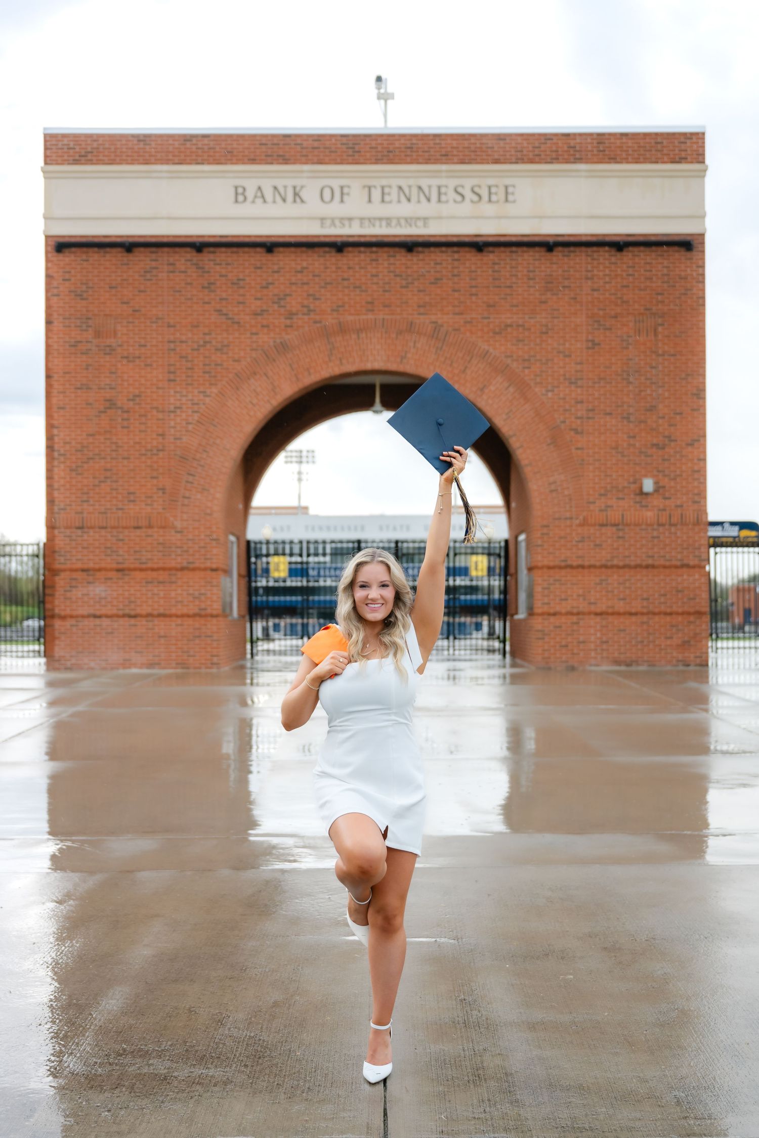 Excited graduate in white dress celebrates with diploma raised at Bank of Tennessee stadium entrance on a rainy day.
