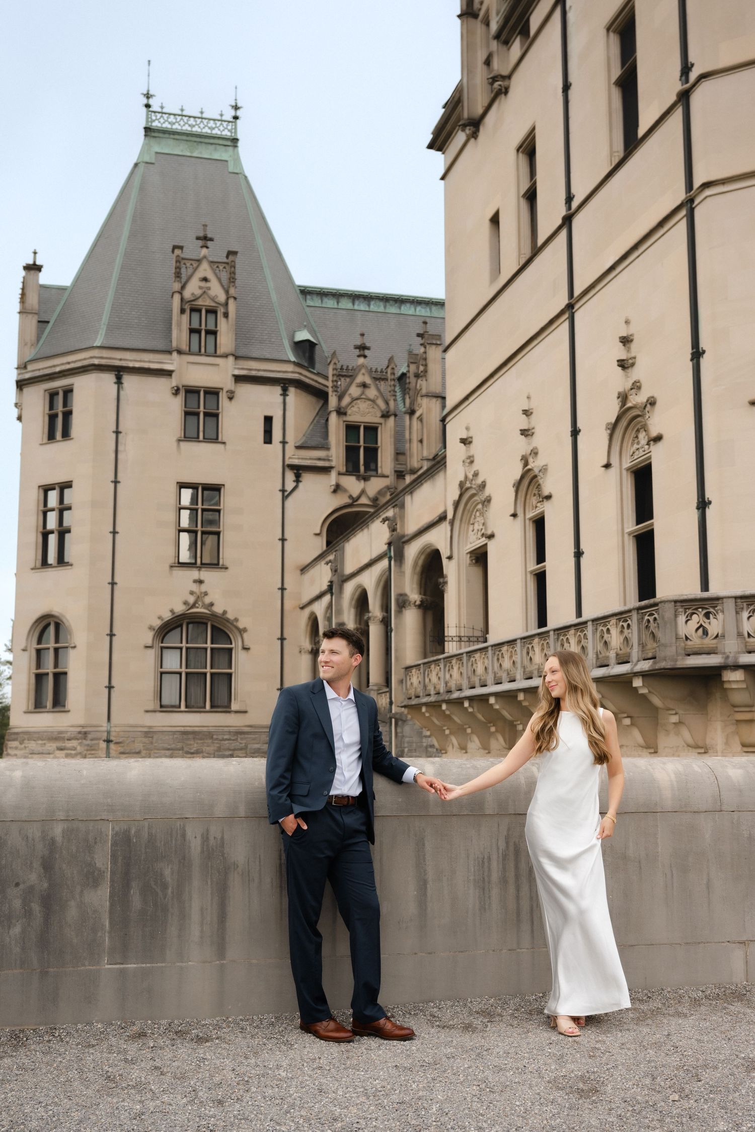 Couple holding hands in front of Biltmore Estate, man in navy suit, woman in white gown, elegant architecture backdrop.