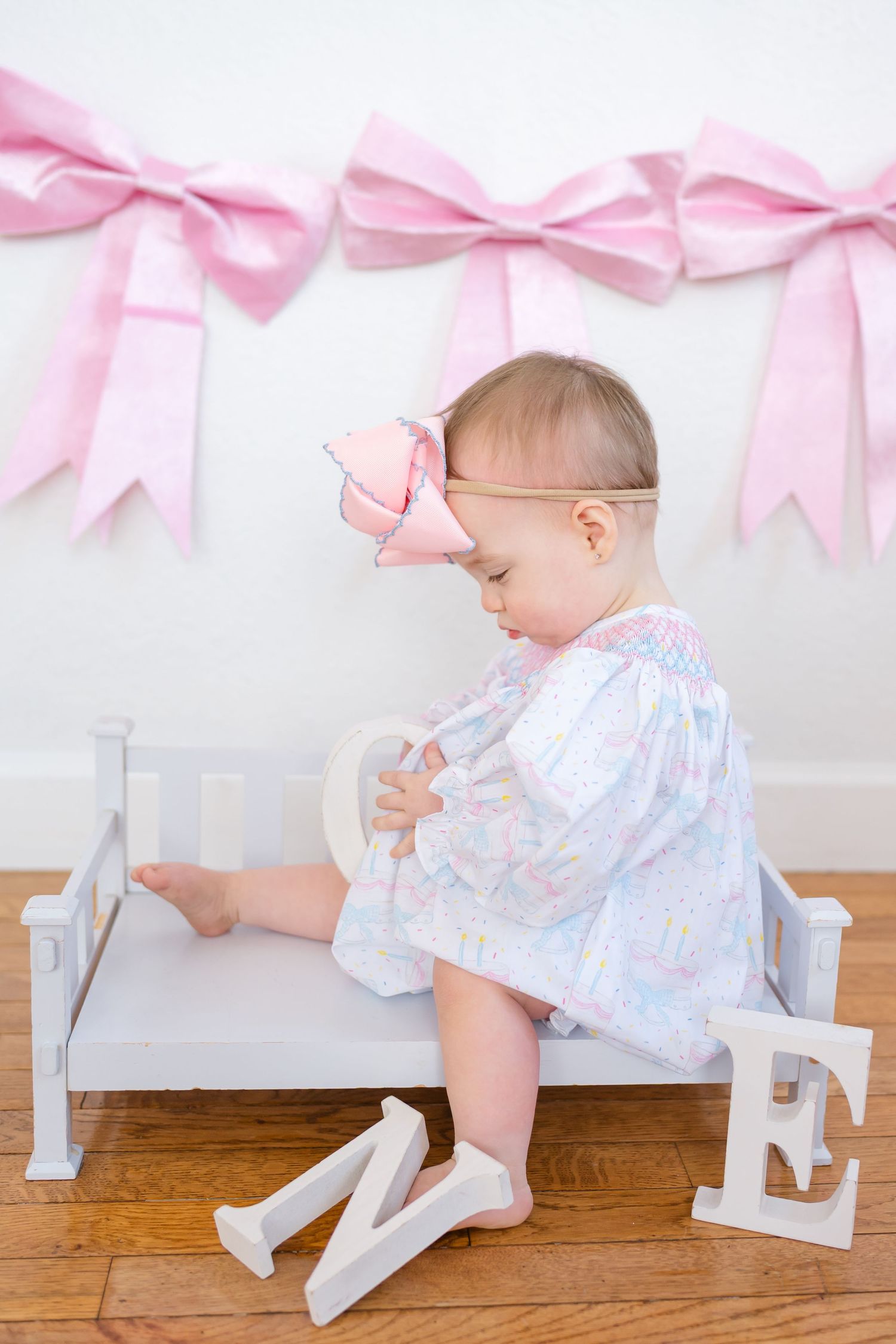 Baby in white dress crawls next to decorative white letters with pink bows hanging above on white wall.