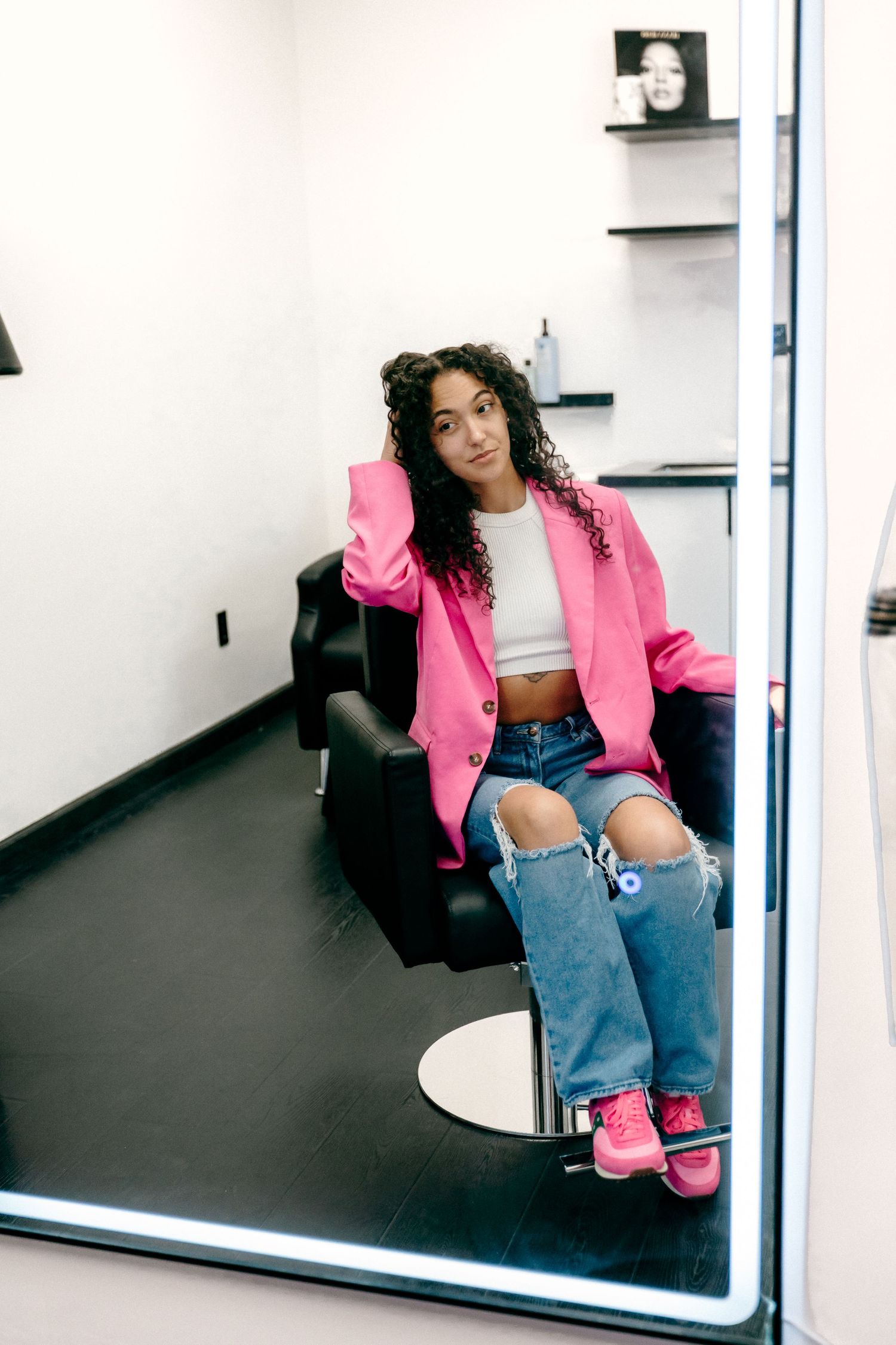 Person in bright pink blazer and ripped jeans poses on black chair in front of mirror.