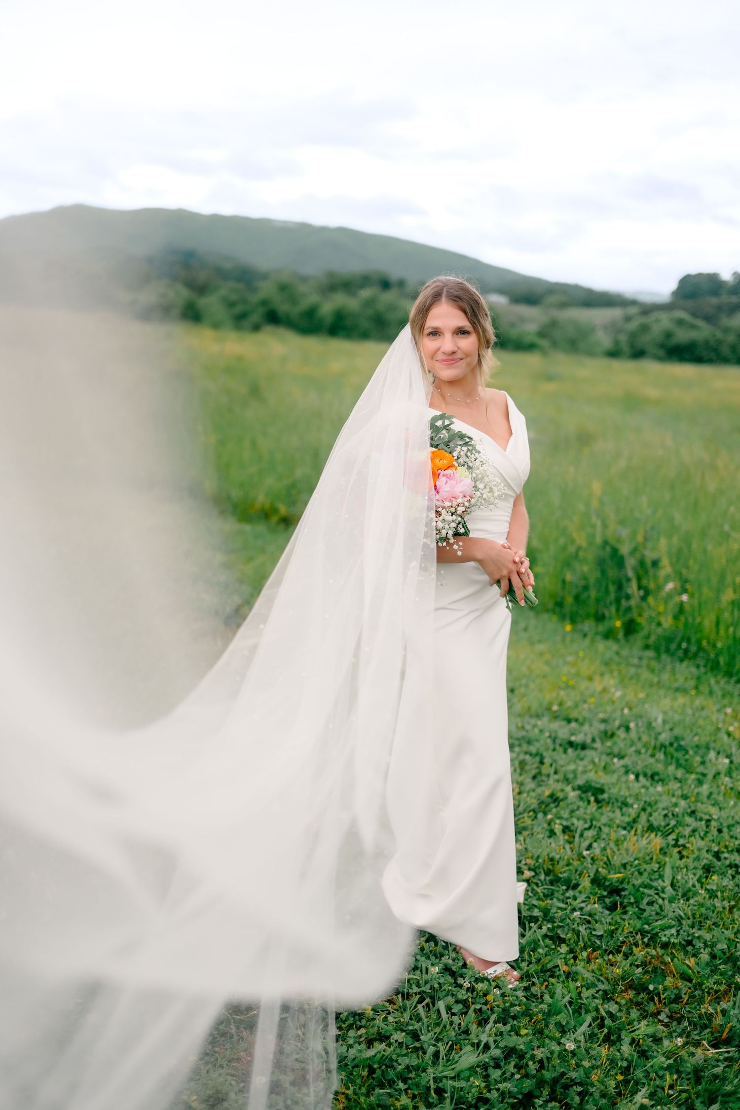 Bride with flowing veil and floral bouquet poses in green countryside.