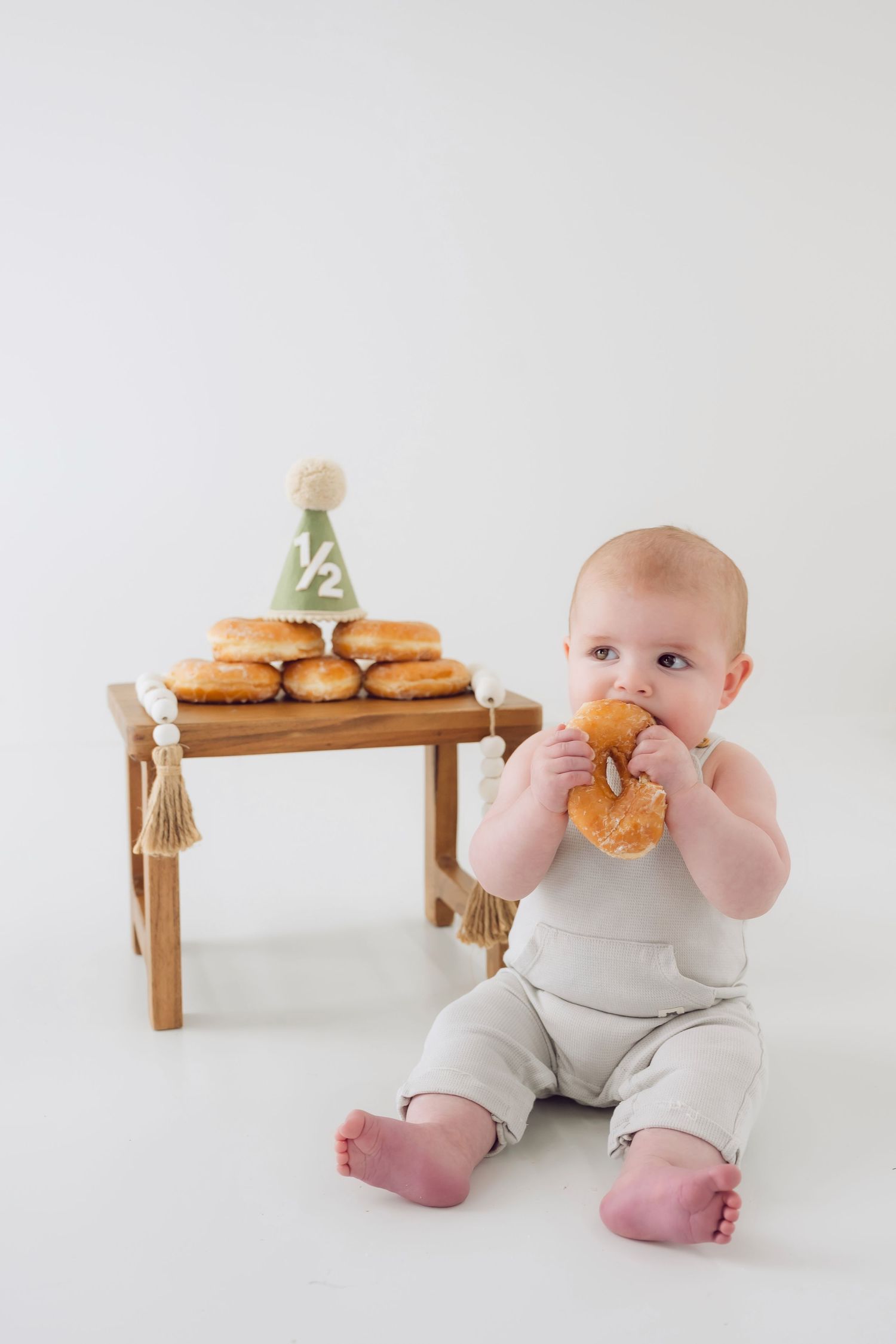 Baby in white romper sits with wooden bench displaying donuts and treats.