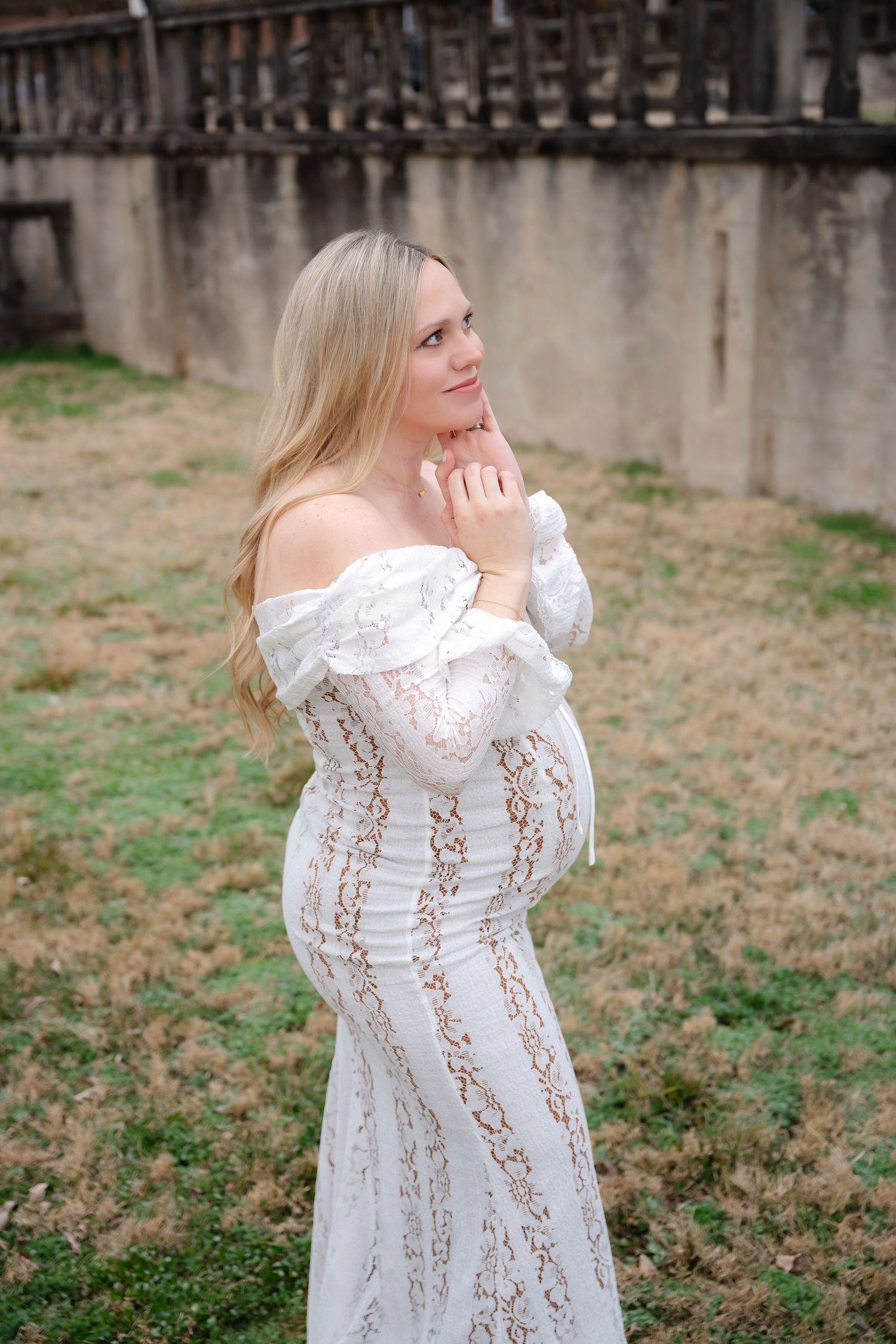 Woman in white lace dress poses gracefully against rustic outdoor backdrop.