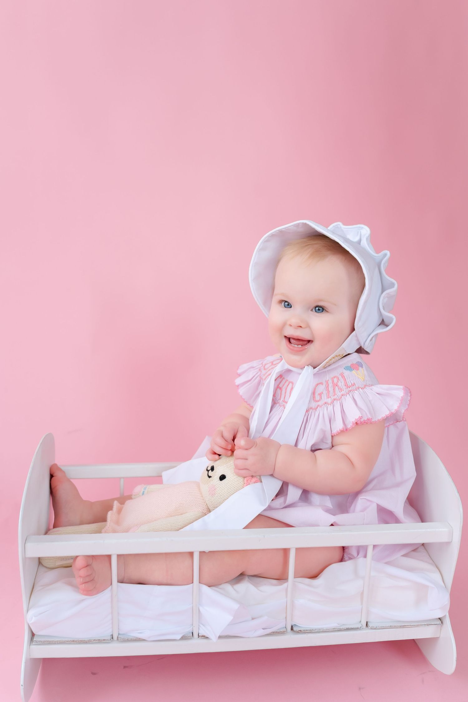 Baby in white dress and bonnet lying on white changing table against pink background in four sequential poses.