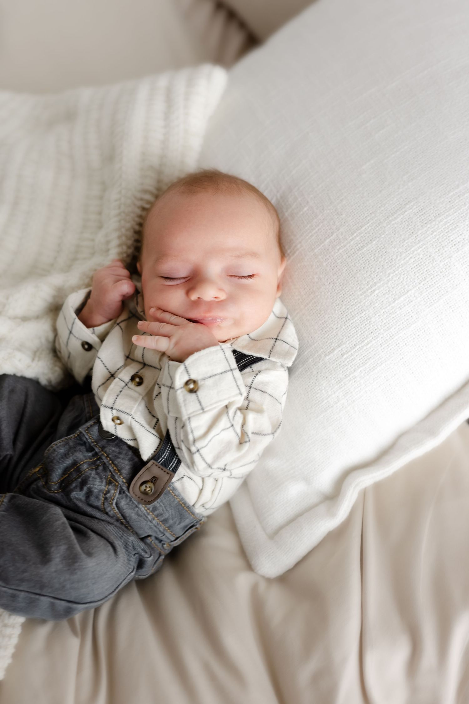 A sleeping baby wearing a white collared shirt and gray pants rests peacefully on white bedding.