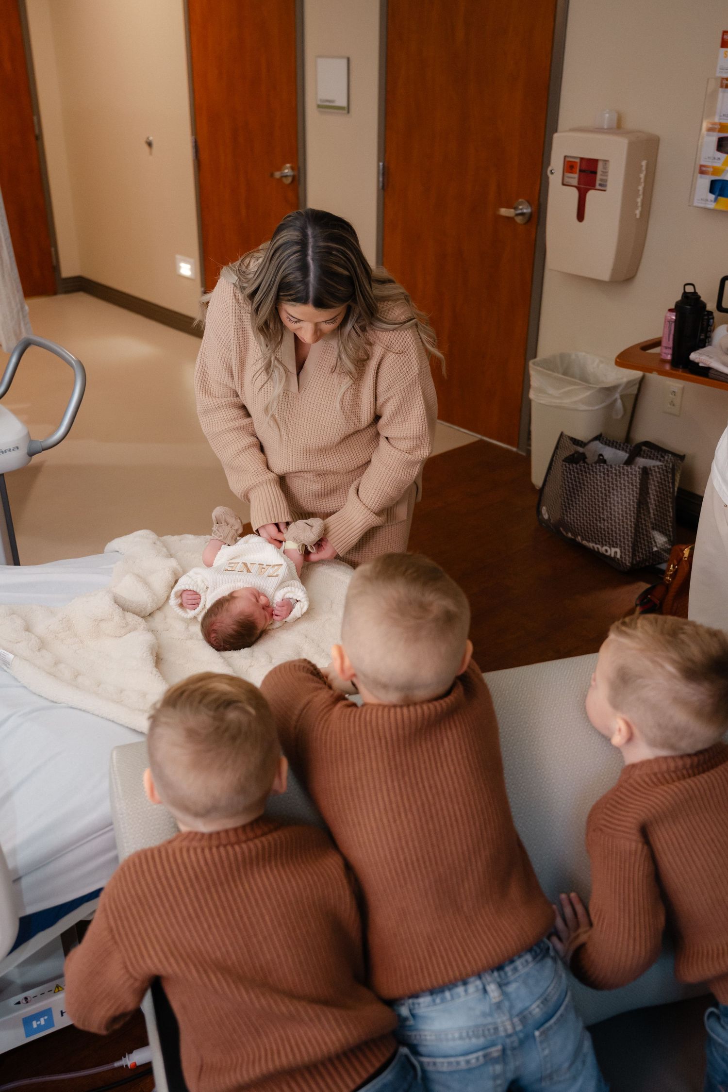 Children in brown sweaters watching through window at hospital nursery.