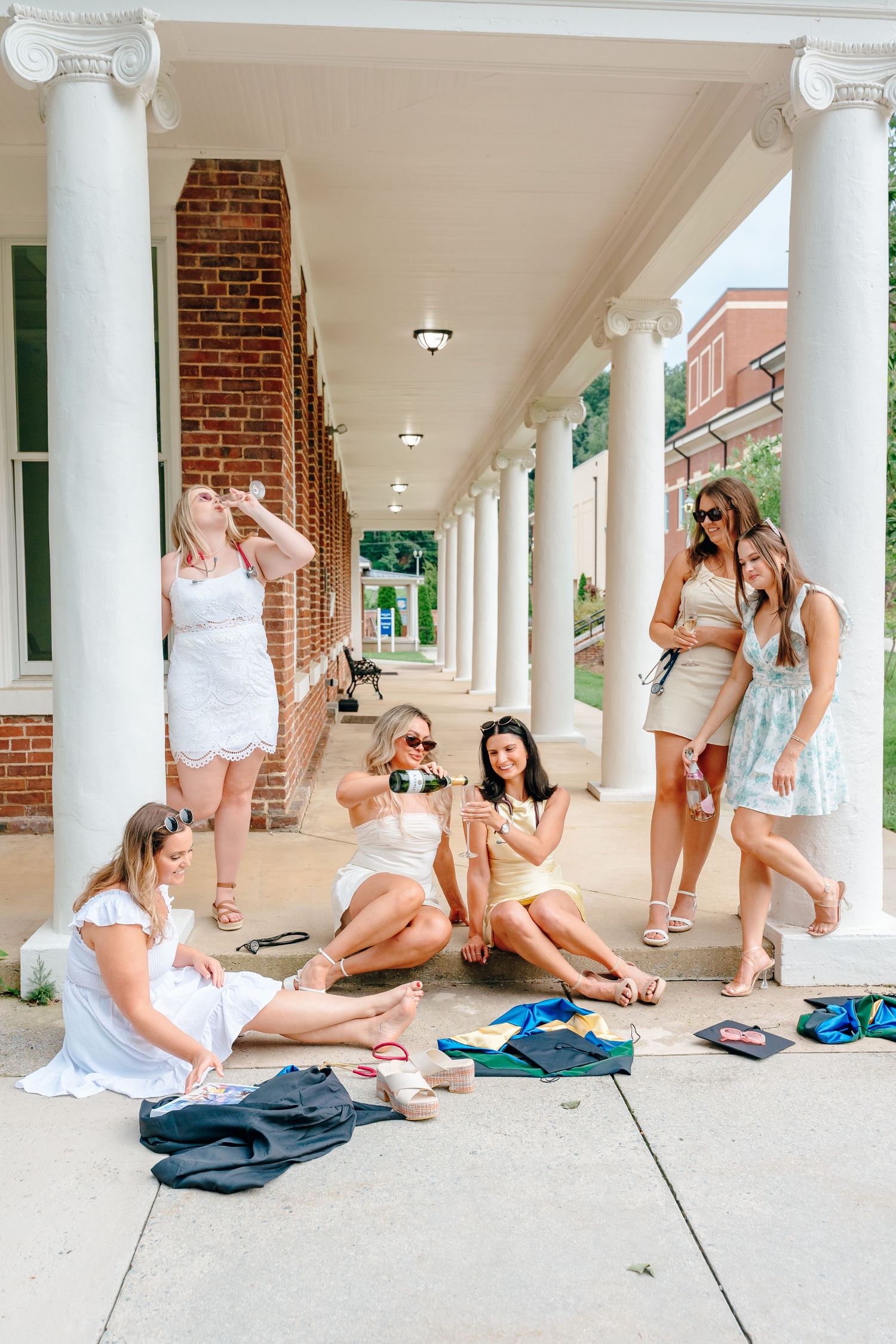 Friends in white outfits sit casually under columned walkway during graduation.