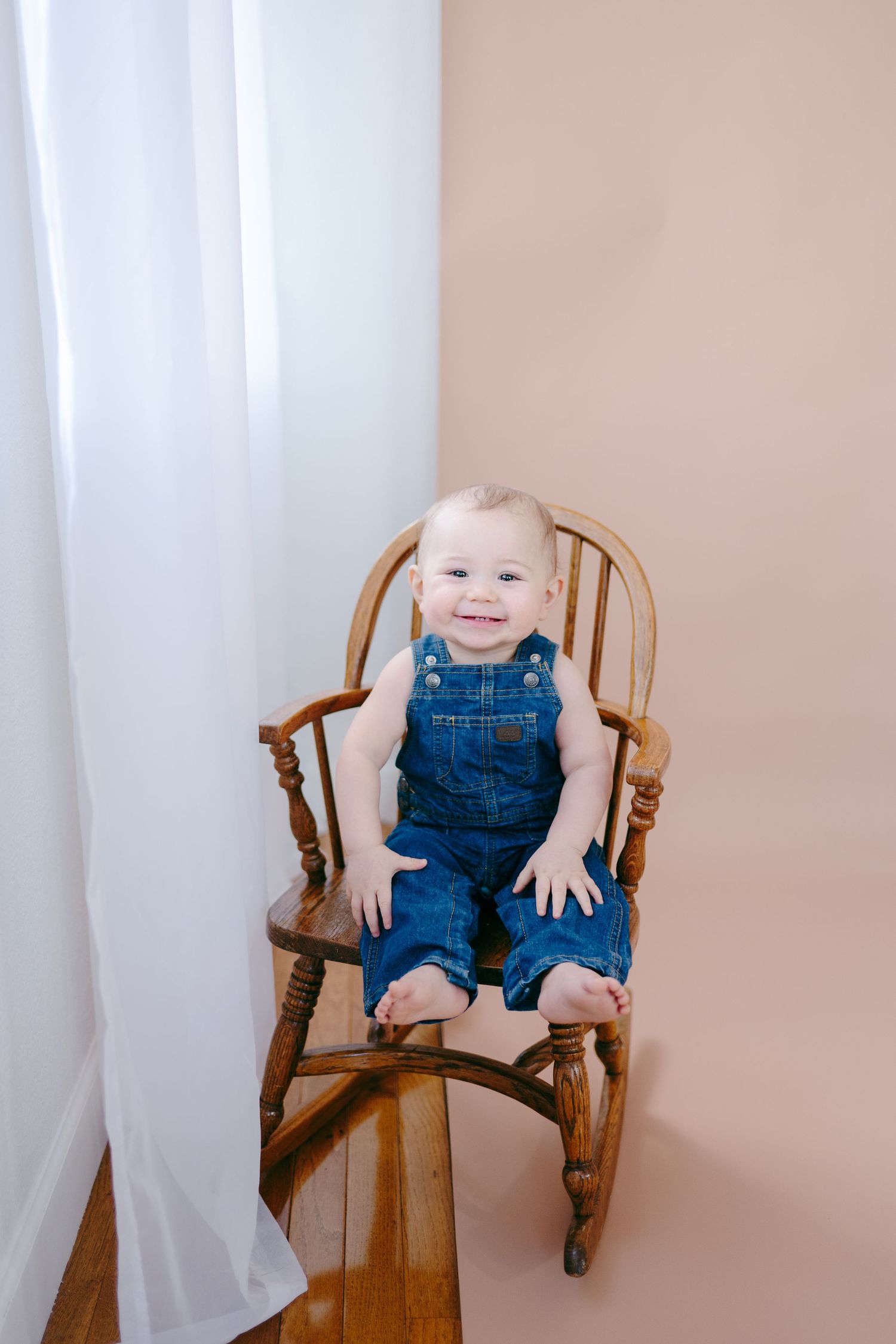 Baby wearing blue overalls sits and smiles in wooden rocking chair.