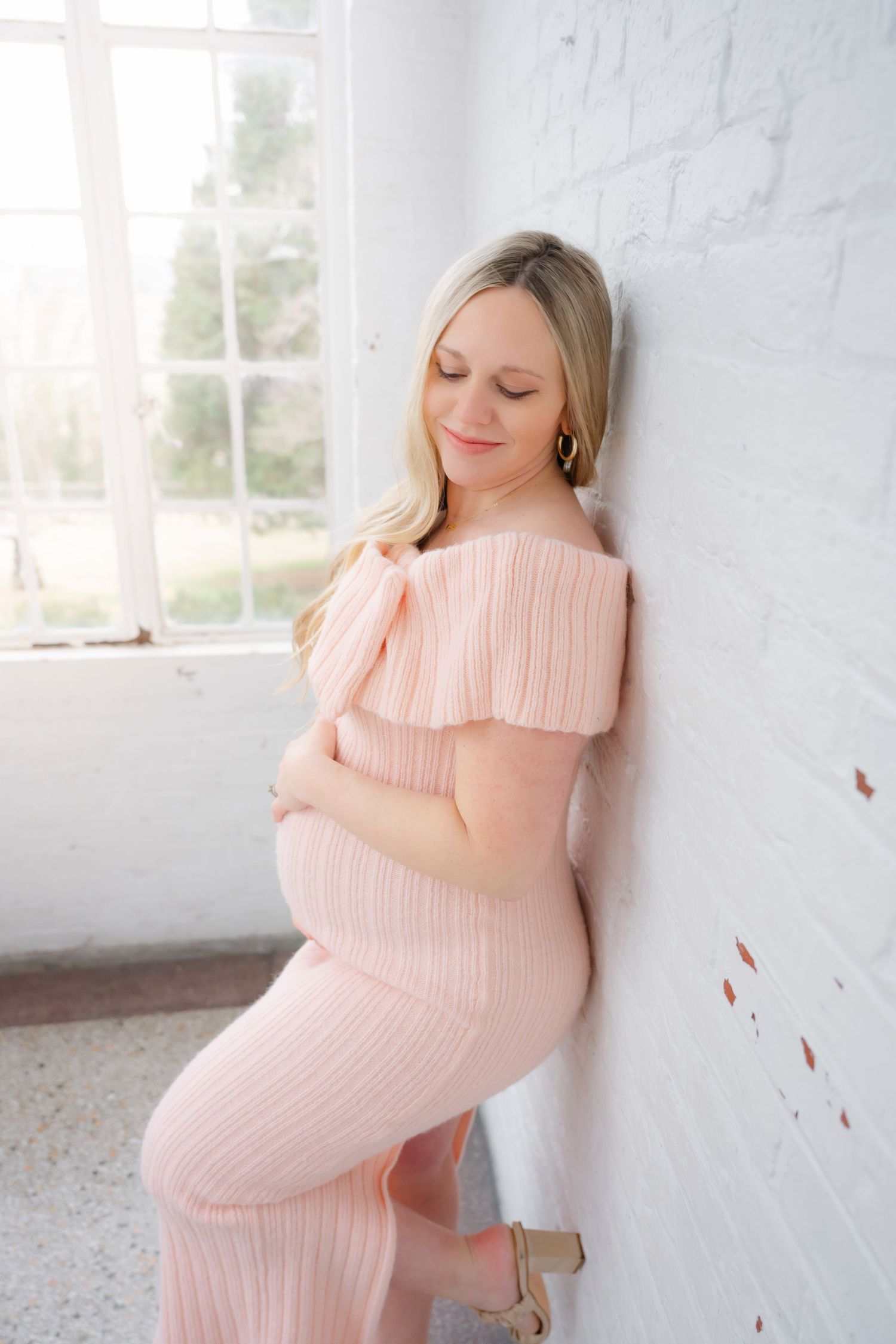 A woman in a pink dress cradles her baby bump against a white wall.